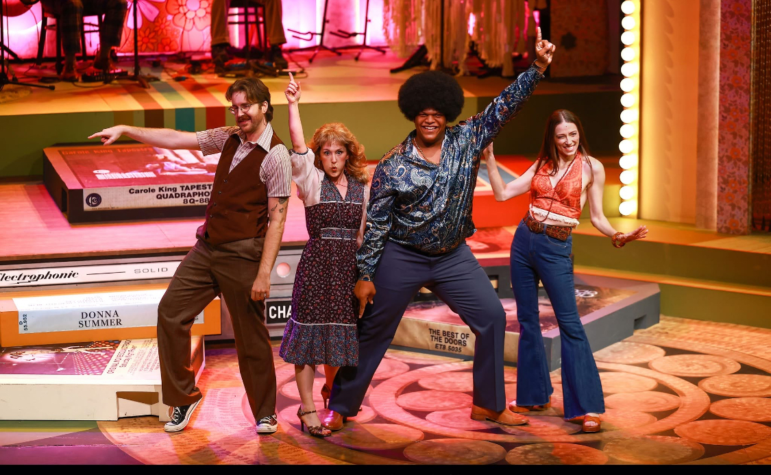 Four people dancing on a colorful stage with books and a keyboard in the background.