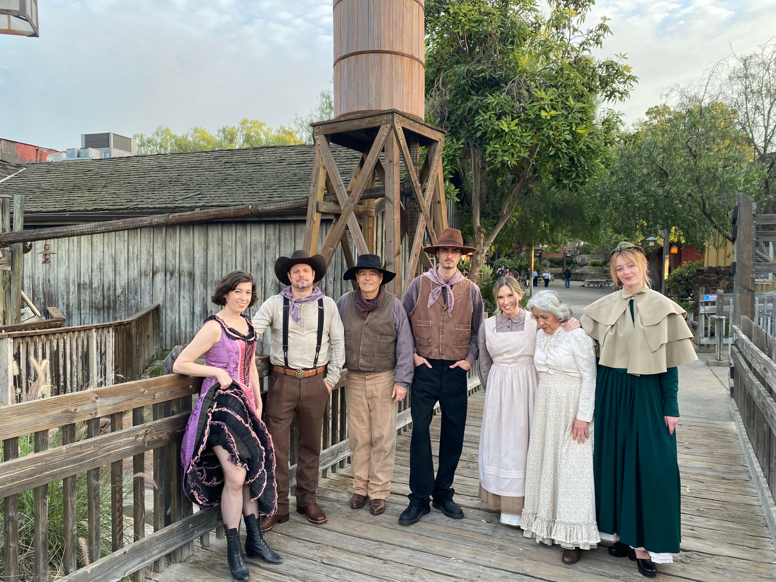 Group of seven people dressed in historical clothing standing on a wooden walkway outdoors, with trees and a wooden structure in the background.
