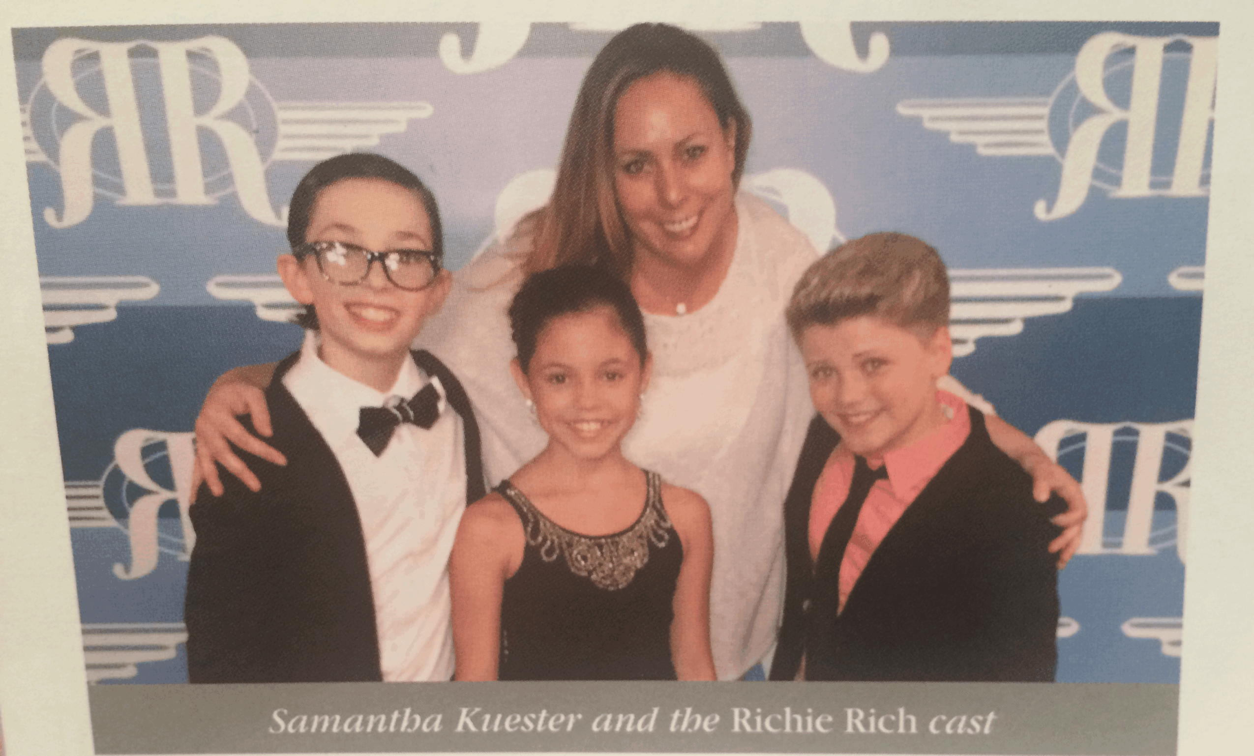 Woman with three children, all smiling, in front of a blue background with white logos, at an event. Text below reads: 'Samantha Kuester and the Richie Rich cast.'