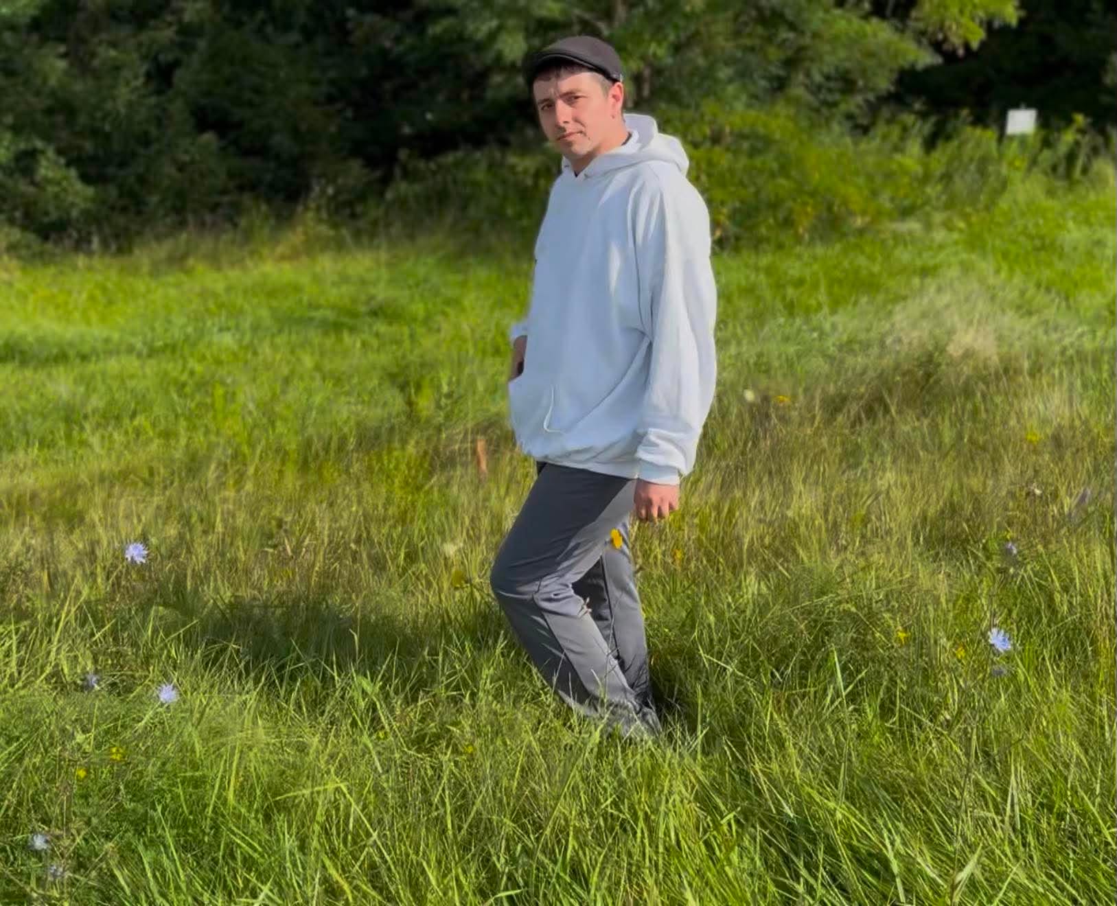 Person standing in a grassy field wearing a light hoodie, dark pants, and a cap, with trees in the background.