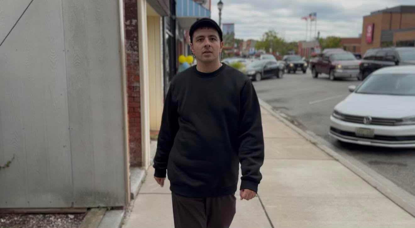 Person walking on sidewalk in urban area, buildings, cars, and street in background.
