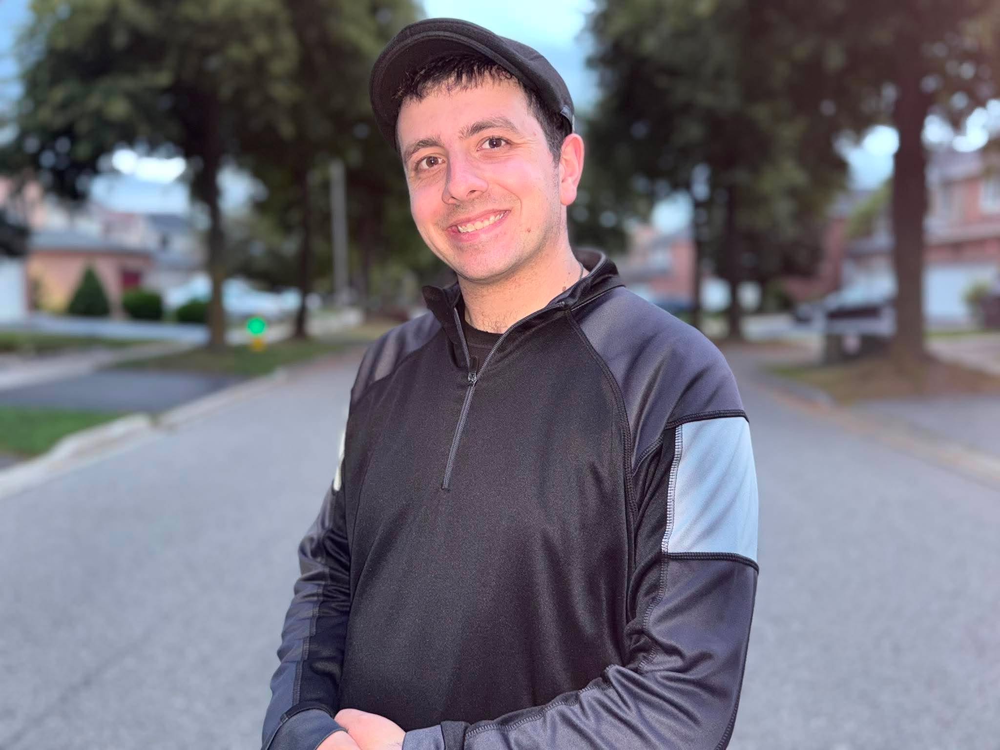 Young man smiling outdoors, wearing a black jacket and cap, standing on a street with trees and houses in background.