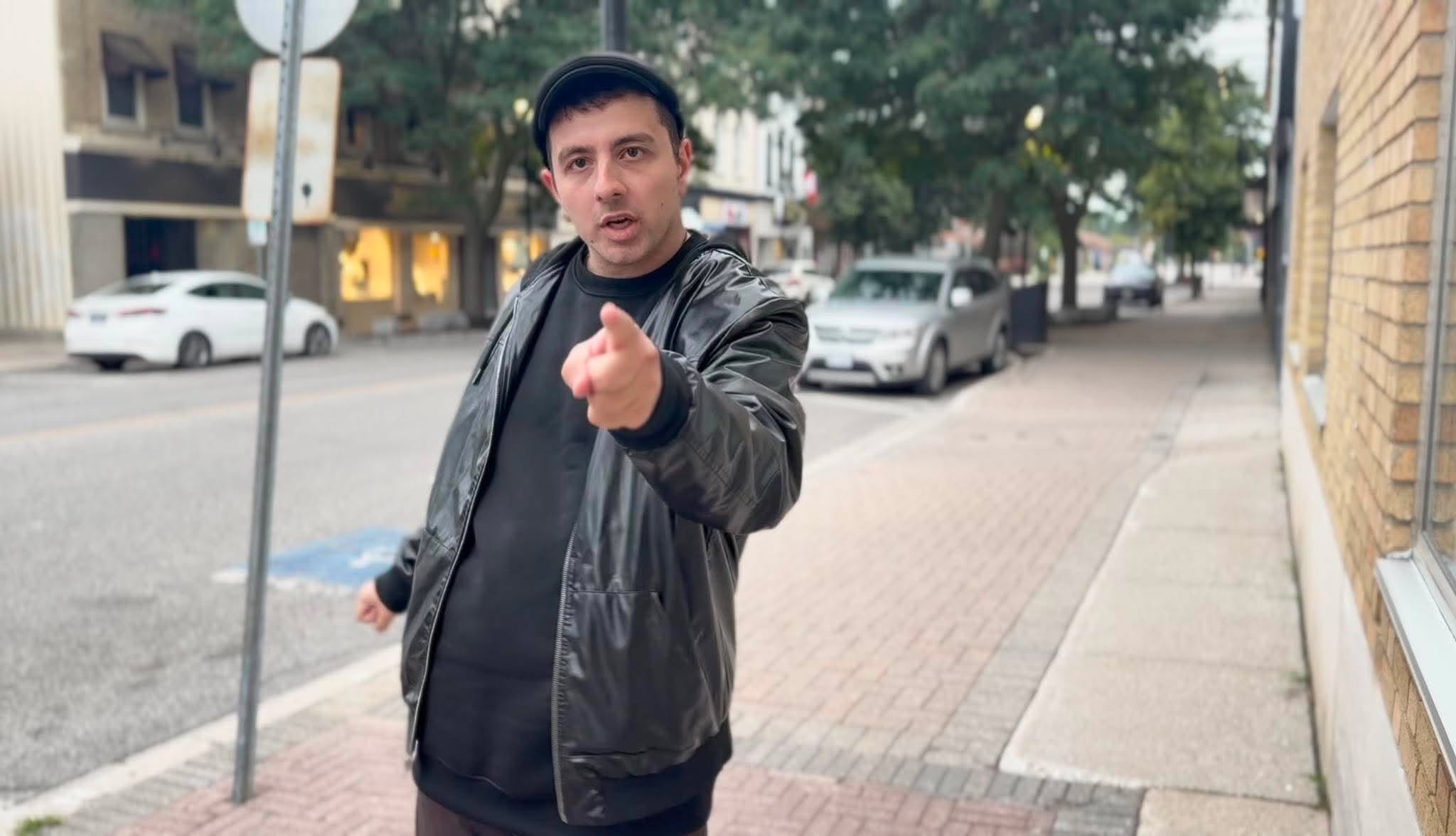 Man in black jacket pointing at camera on sidewalk with trees and parked cars in background