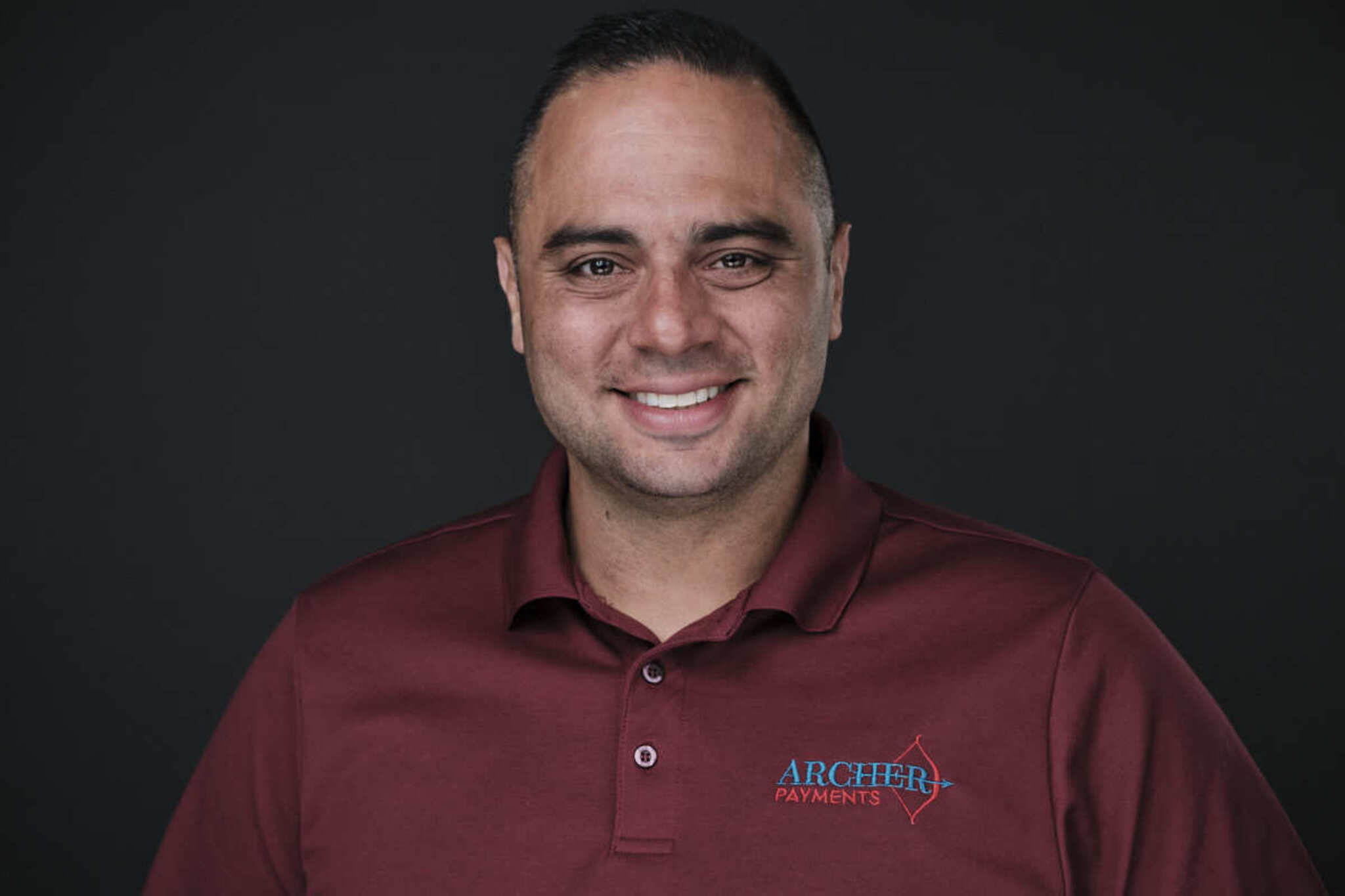 Man with short dark hair and a beard, smiling, wearing a maroon polo shirt with a logo, against a dark background.