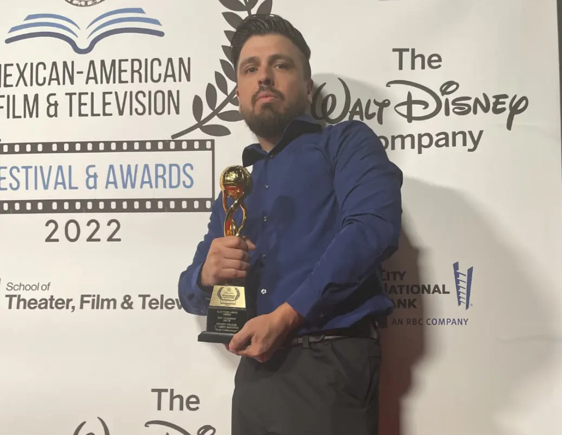 Man holding an award trophy in front of a backdrop with logos and text, wearing a blue shirt and dark pants.
