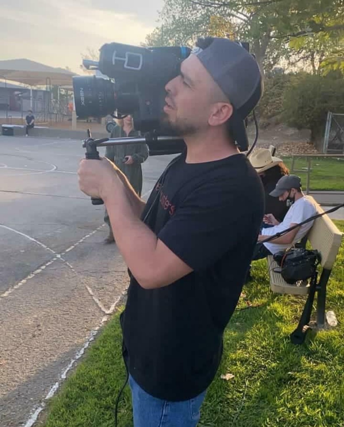 Young man with a cap holding a professional camera on a stabilizer outdoors, with a person sitting on a bench in the background.