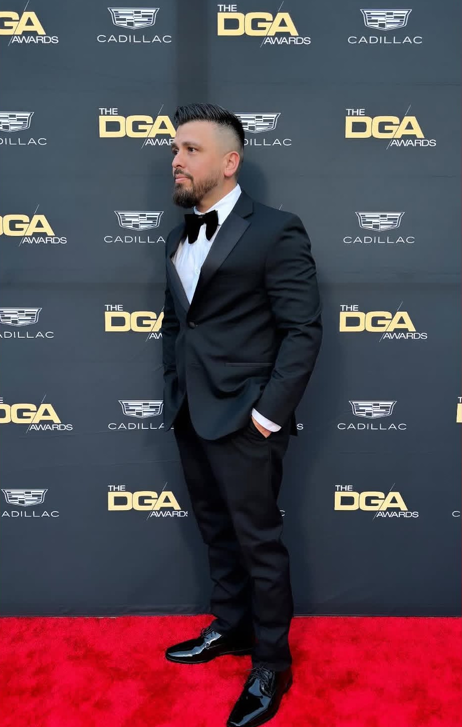 Man in tuxedo with bow tie standing on red carpet against backdrop with logos, facing right, hands in pockets.