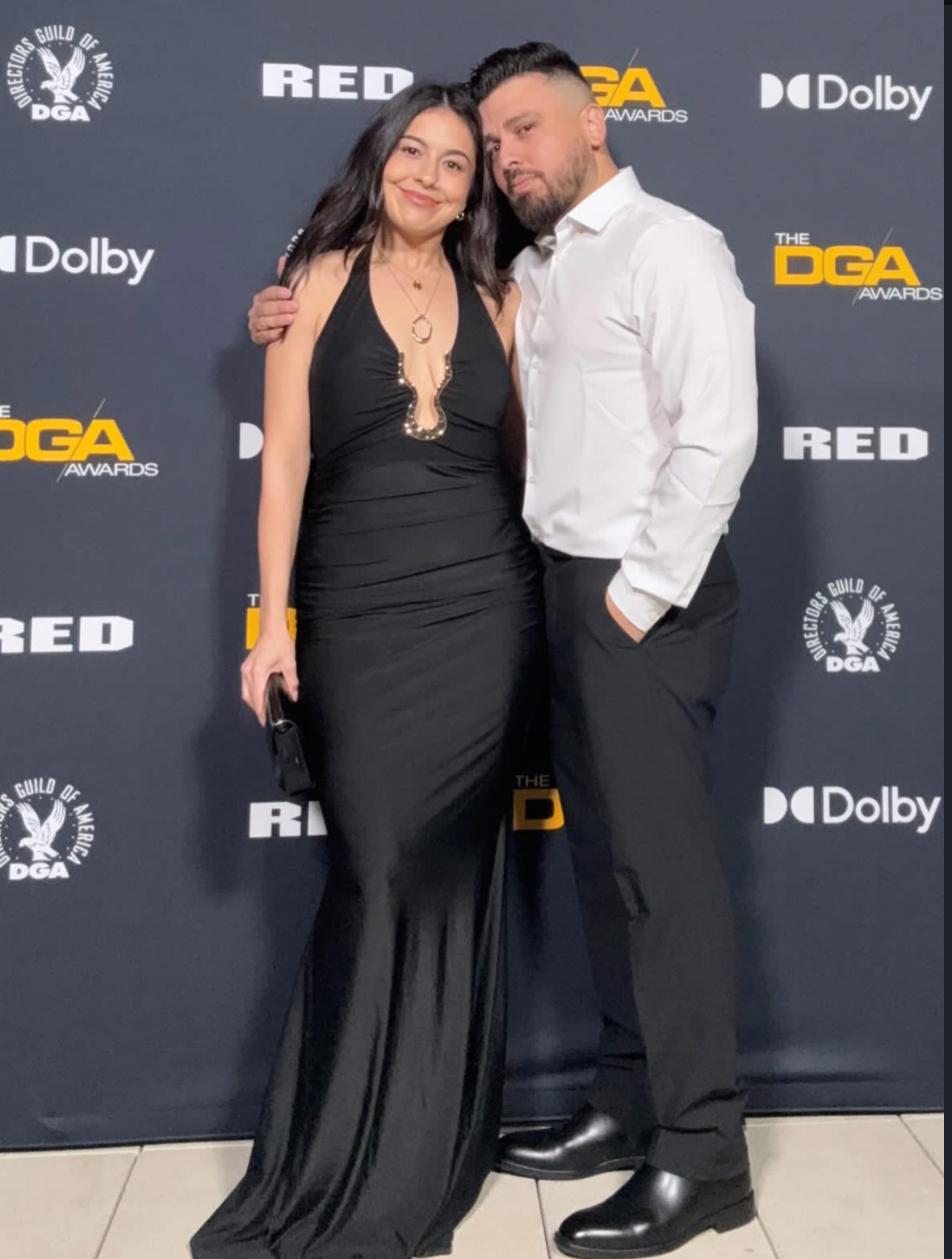 A woman in a black gown and a man in a white shirt and dark pants pose together on a red carpet backdrop.