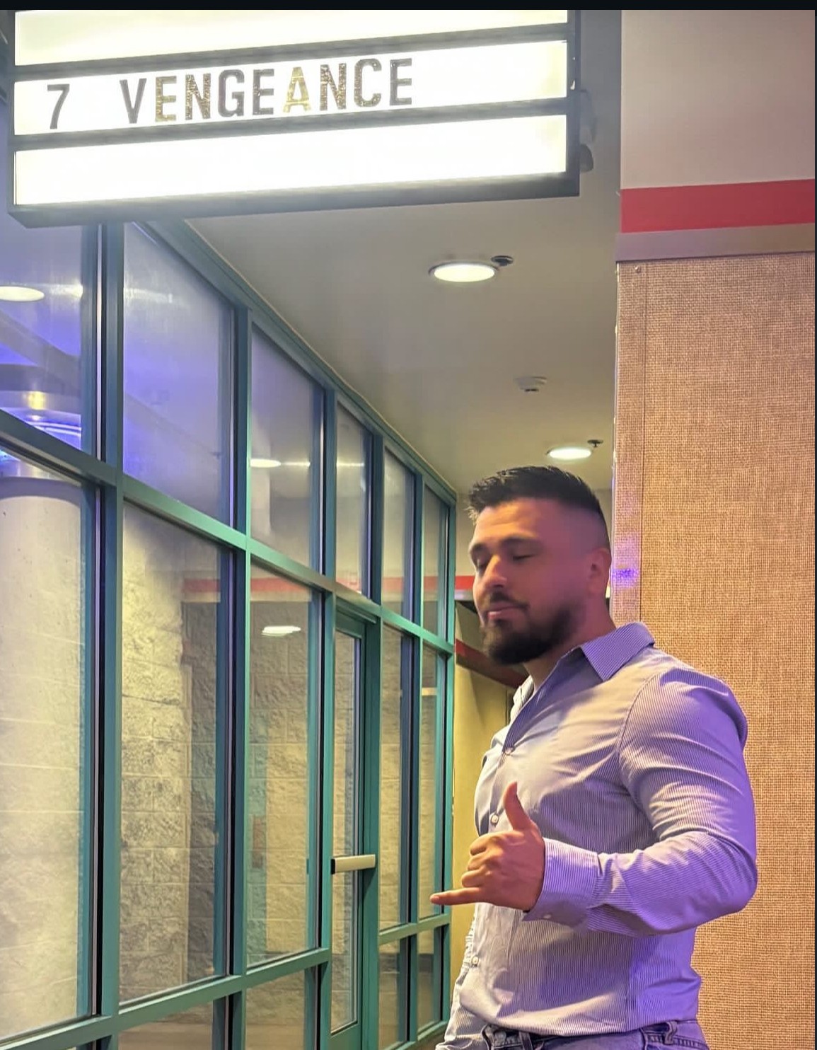 Man with beard and short hair in a light purple shirt making a hand gesture, standing indoors near glass windows.