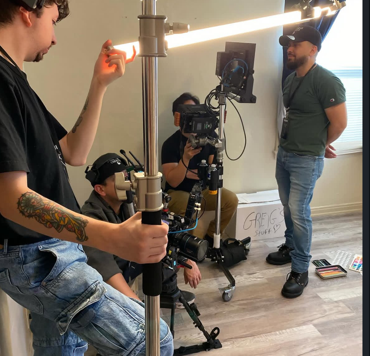Group of people filming a scene indoors with camera equipment and lighting, one person standing, others crouching or sitting.