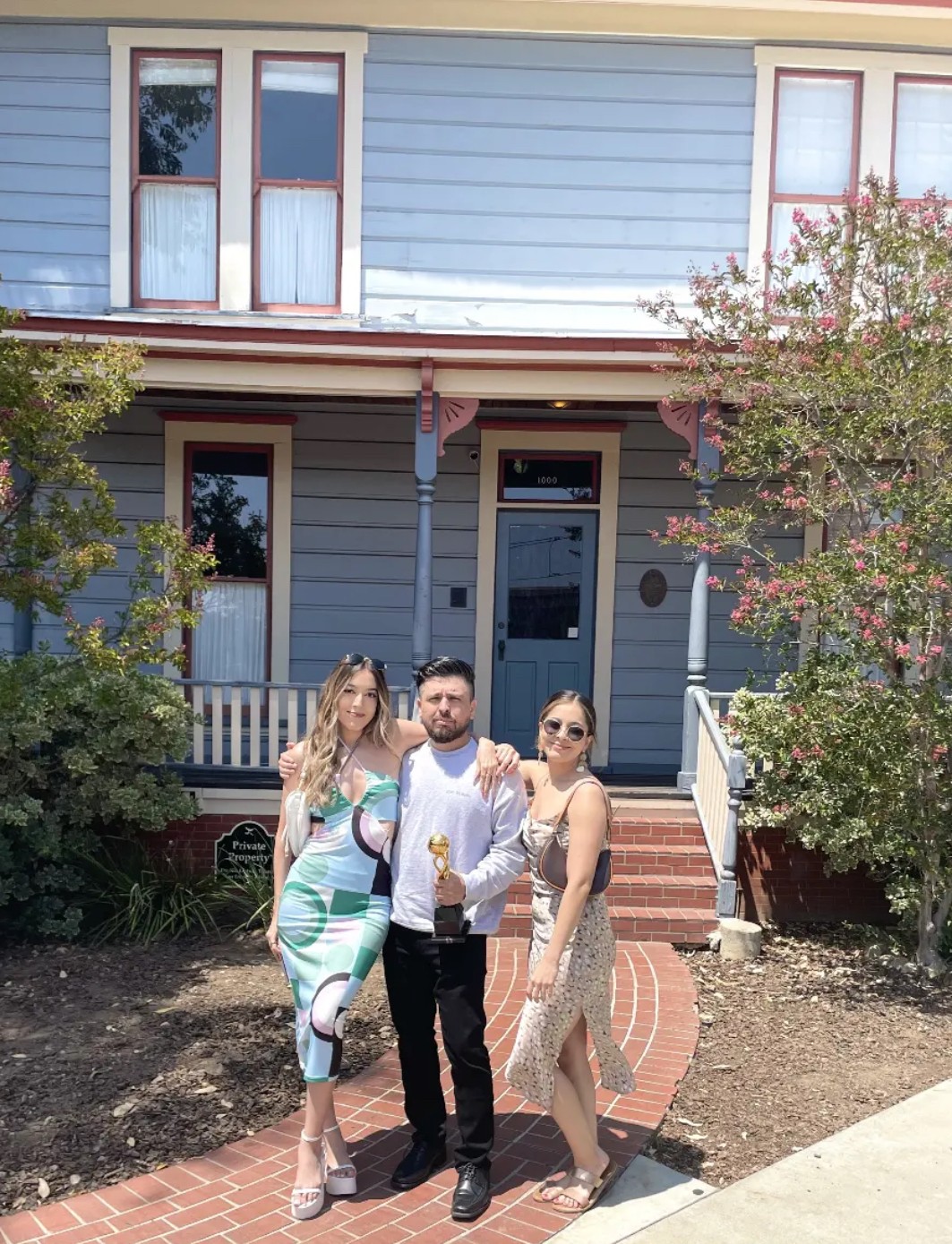 Three people standing in front of a house with blue siding, trees, and a brick pathway, smiling at the camera.