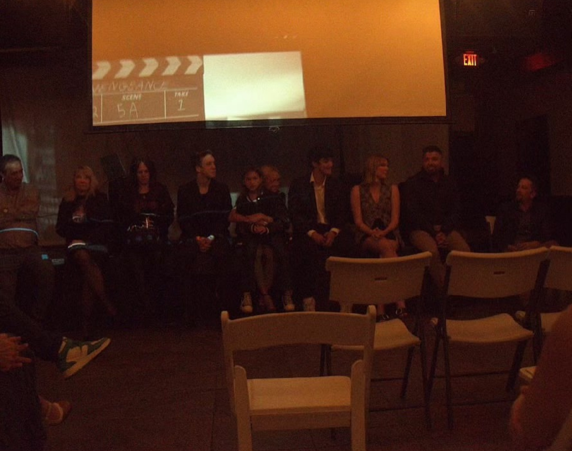 People sitting on a stage in a dark room with a large screen behind them, some chairs in front, dim lighting.