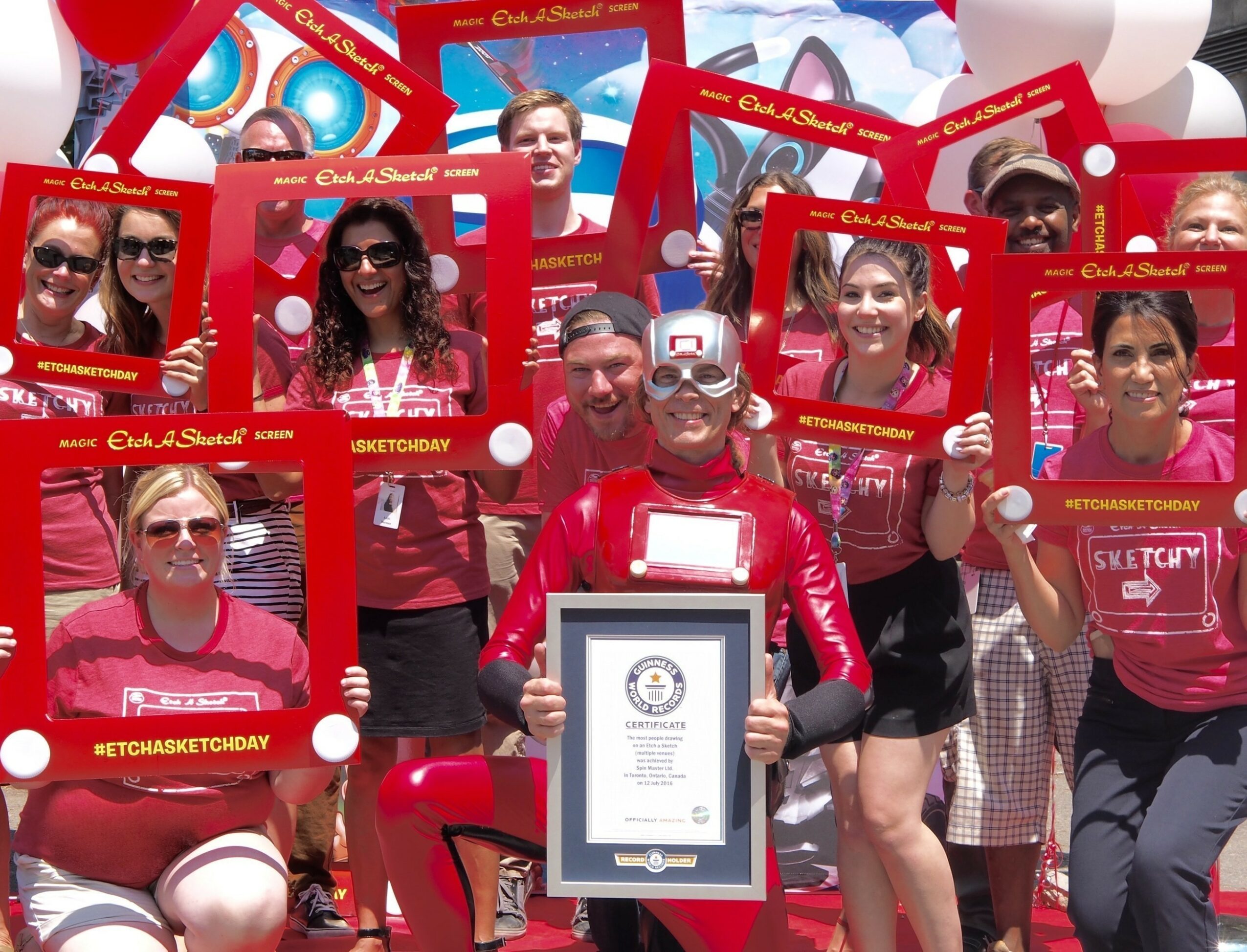 Group of people holding red photo frames, celebrating outdoors with balloons and a large check, smiling at camera.