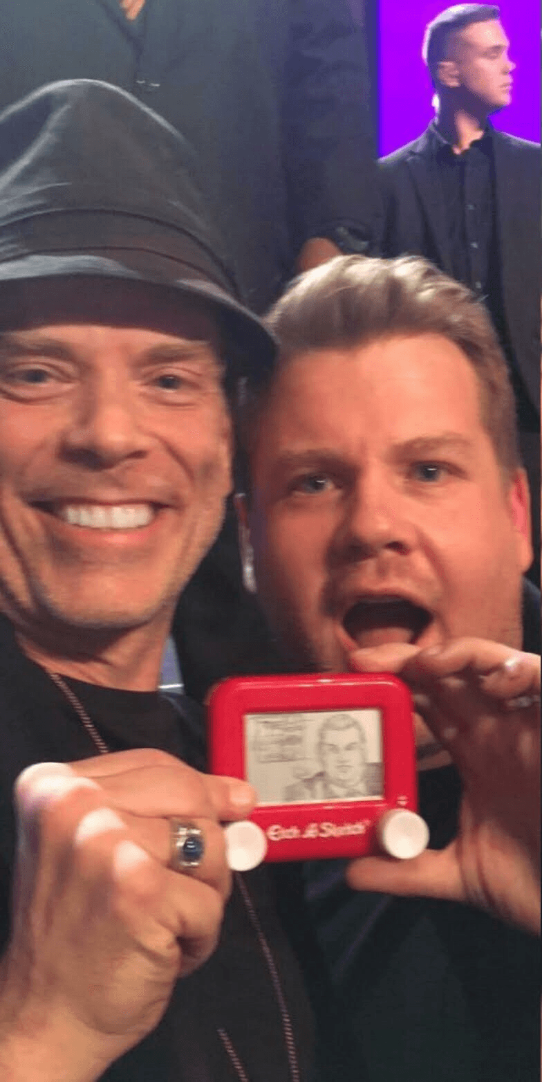 Two men smiling, one holding a red device with a photo, in a crowd with a purple background.