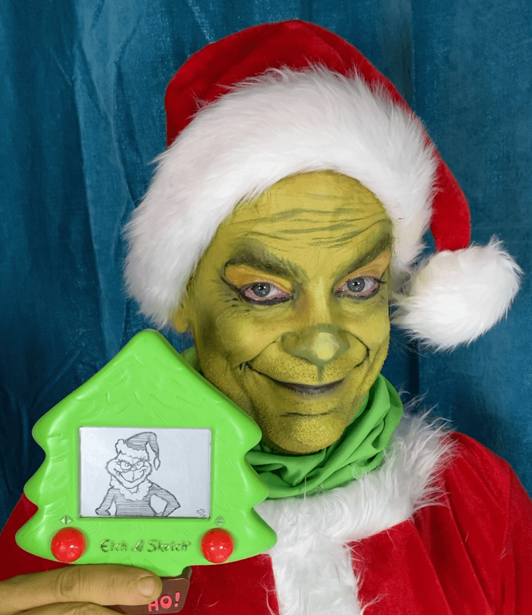 Person dressed as the Grinch wearing a Santa hat and red costume, holding a green drawing tablet, smiling.