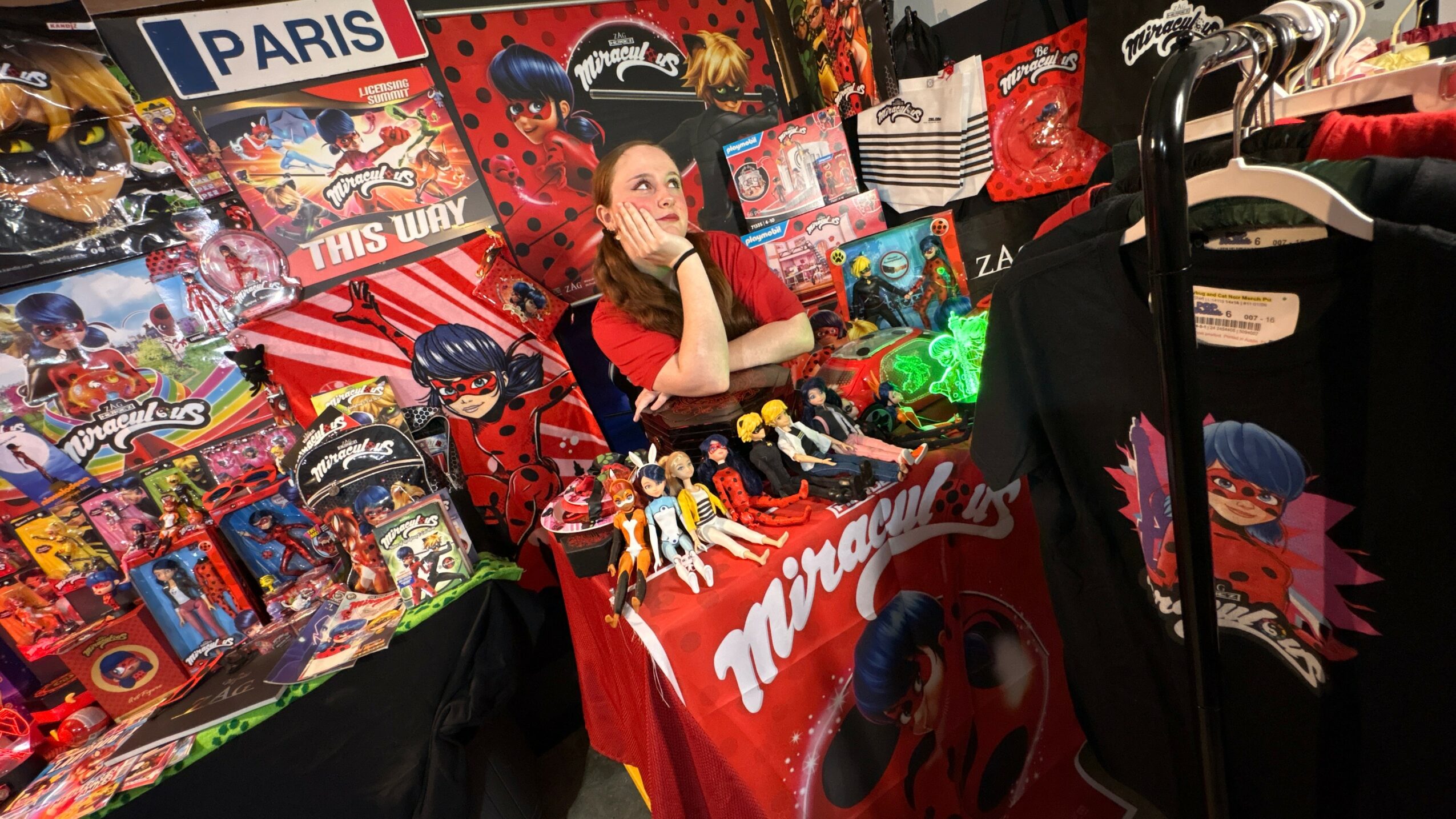 A woman sits at a table with Marvel and superhero merchandise, surrounded by toys, posters, and clothing, in a colorful display.