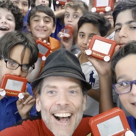 Group of children and a man taking a selfie, holding red electronic devices, smiling and looking at the camera.