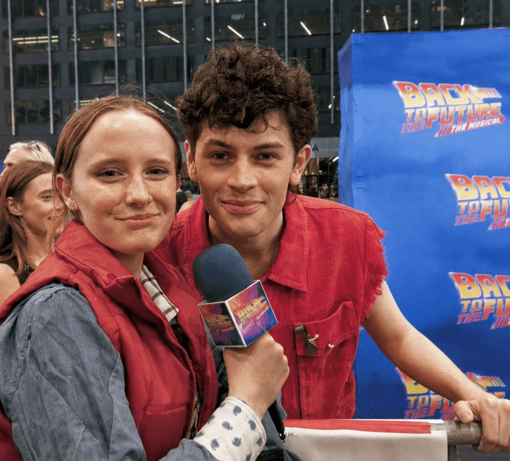 Two young people, one holding a microphone, pose for a photo at an event with a blue backdrop.