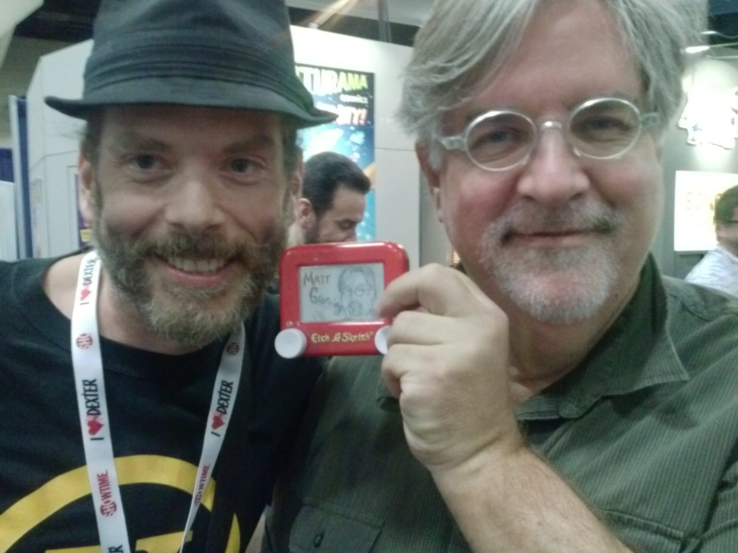 Two men smiling, one holding a red device, in an indoor setting with people and displays in the background.