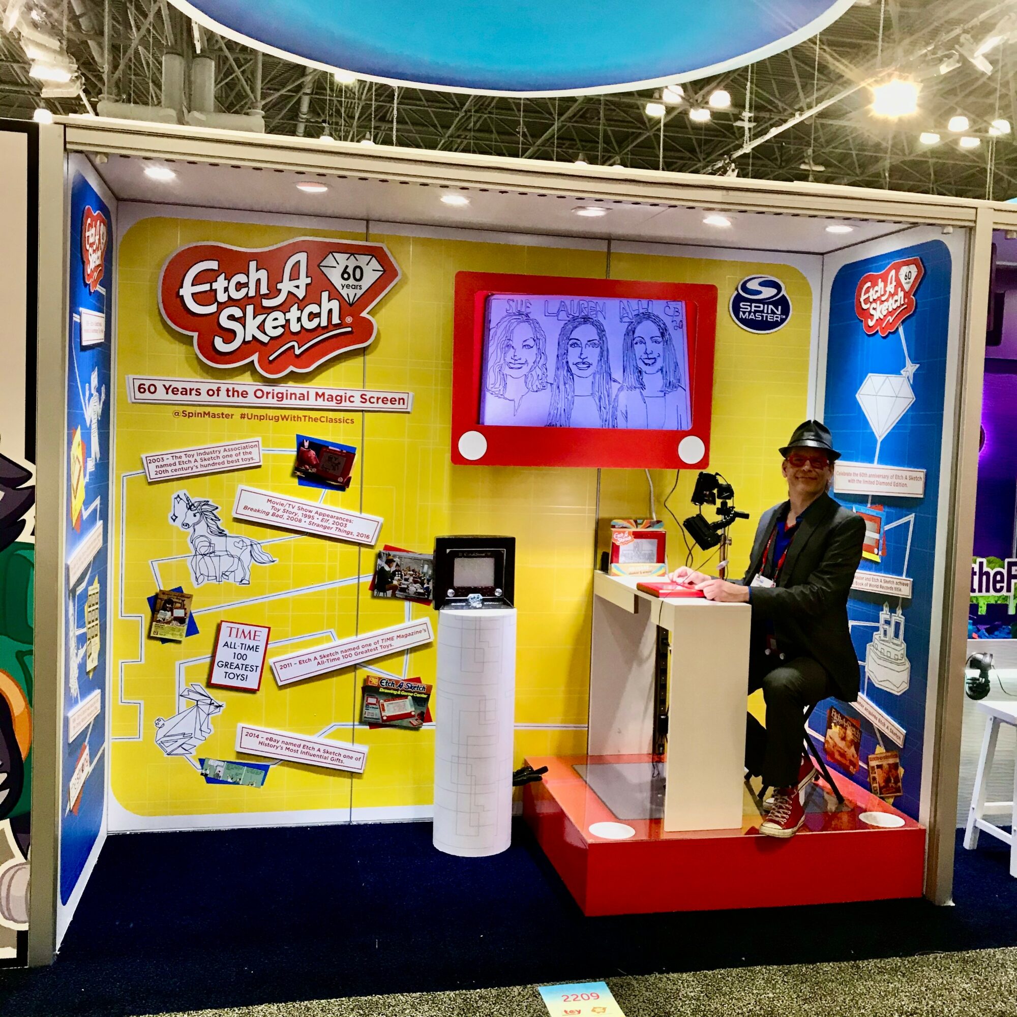 Person sitting at Etch A Sketch booth with drawing on screen, colorful display, and promotional materials, celebrating 60 years of the original magic screen.