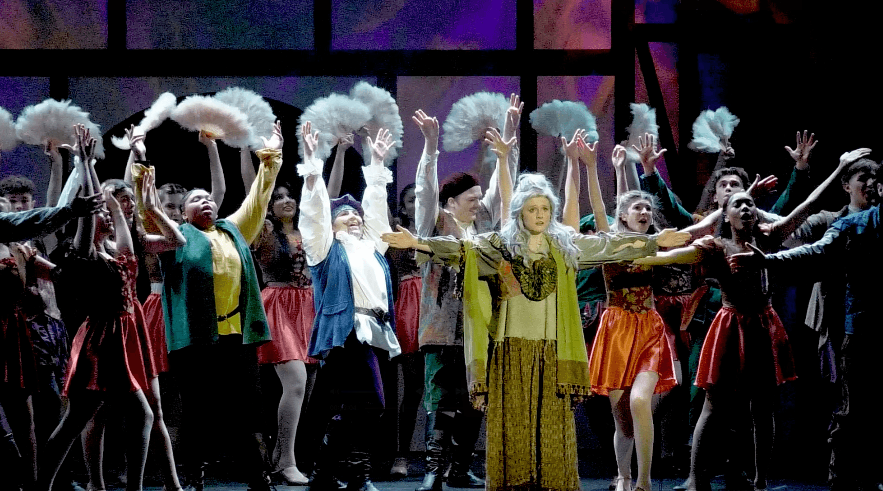 Group of performers on stage holding white fans, with a woman in a yellow dress in the center, stage lighting visible.