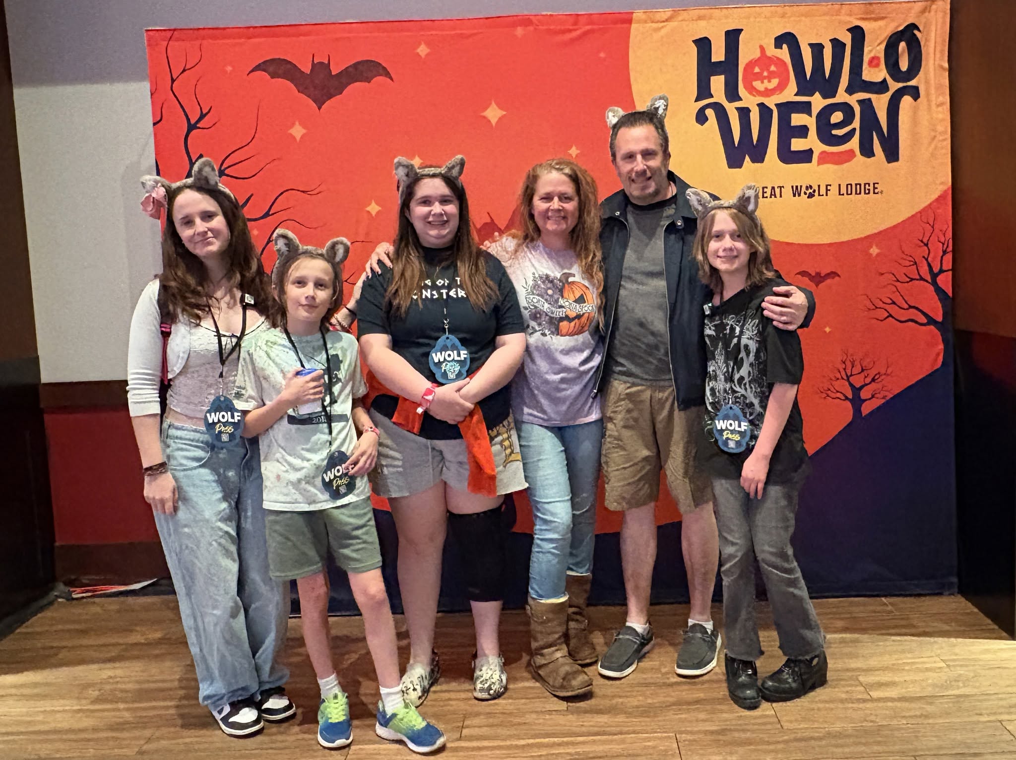 Group of six people standing in front of a Halloween-themed backdrop with a red sky, bats, and trees, smiling at the camera.