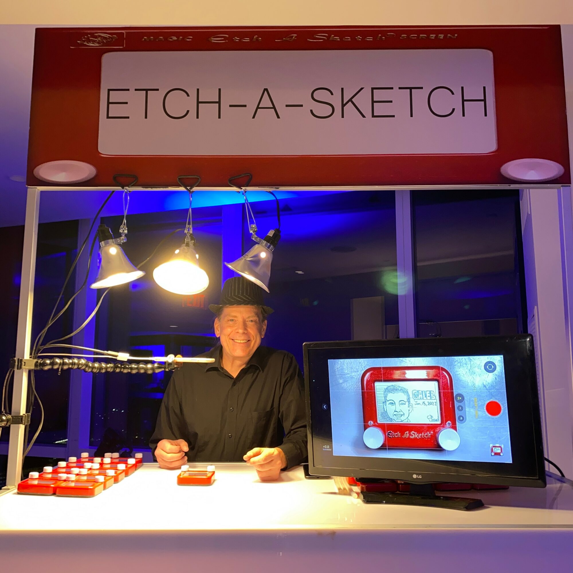Person standing behind a table with a sign reading 'ETCH-A-SKETCH', illuminated by hanging lights, with a screen displaying an Etch-A-Sketch image.