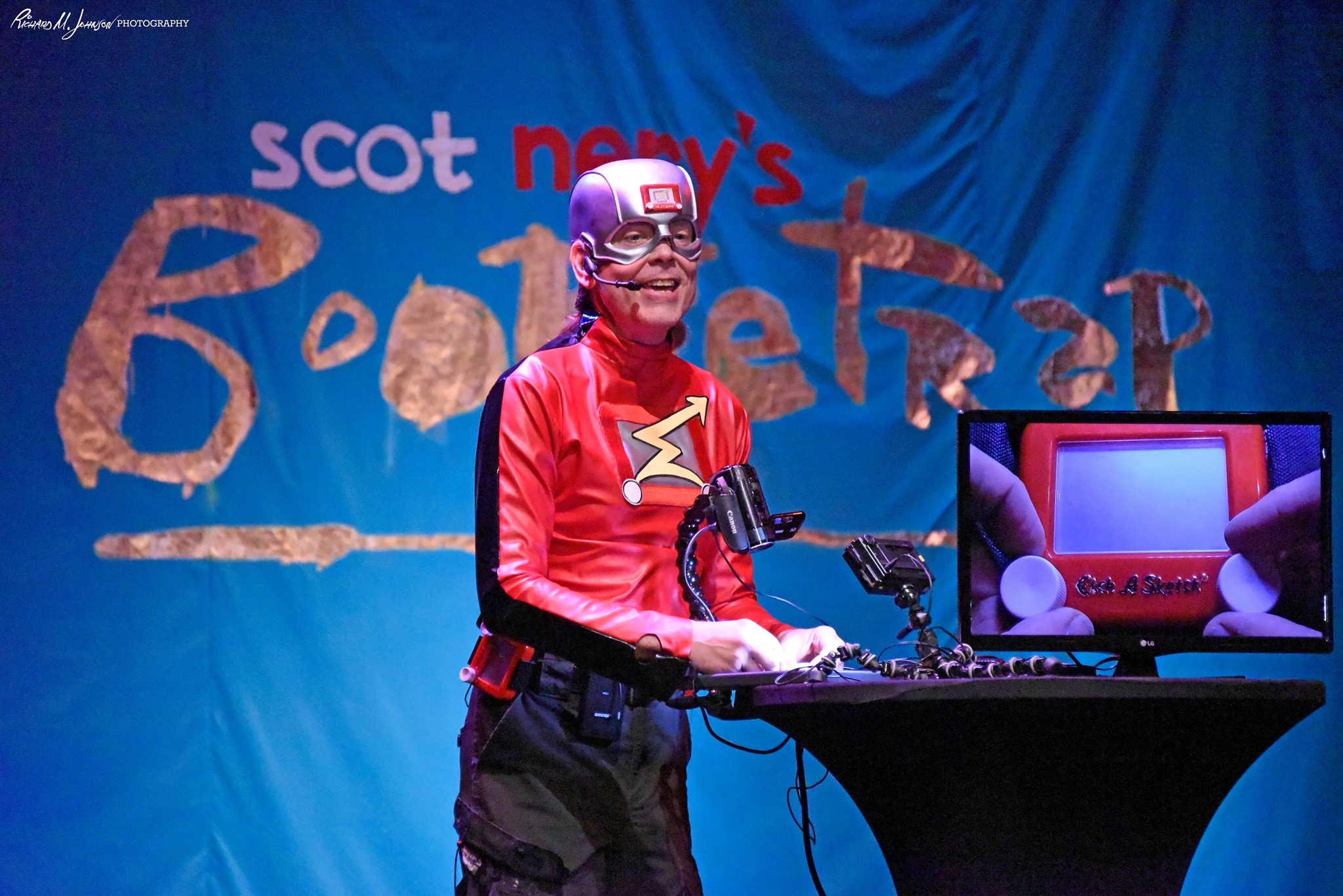 Person dressed as a superhero with a red costume and mask, standing at a table with a monitor, in front of a blue backdrop.