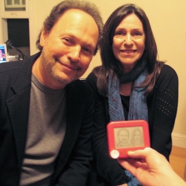 Two people smiling indoors, one holding a small red electronic device with a screen showing two faces, in a room with a window and a computer in the background.