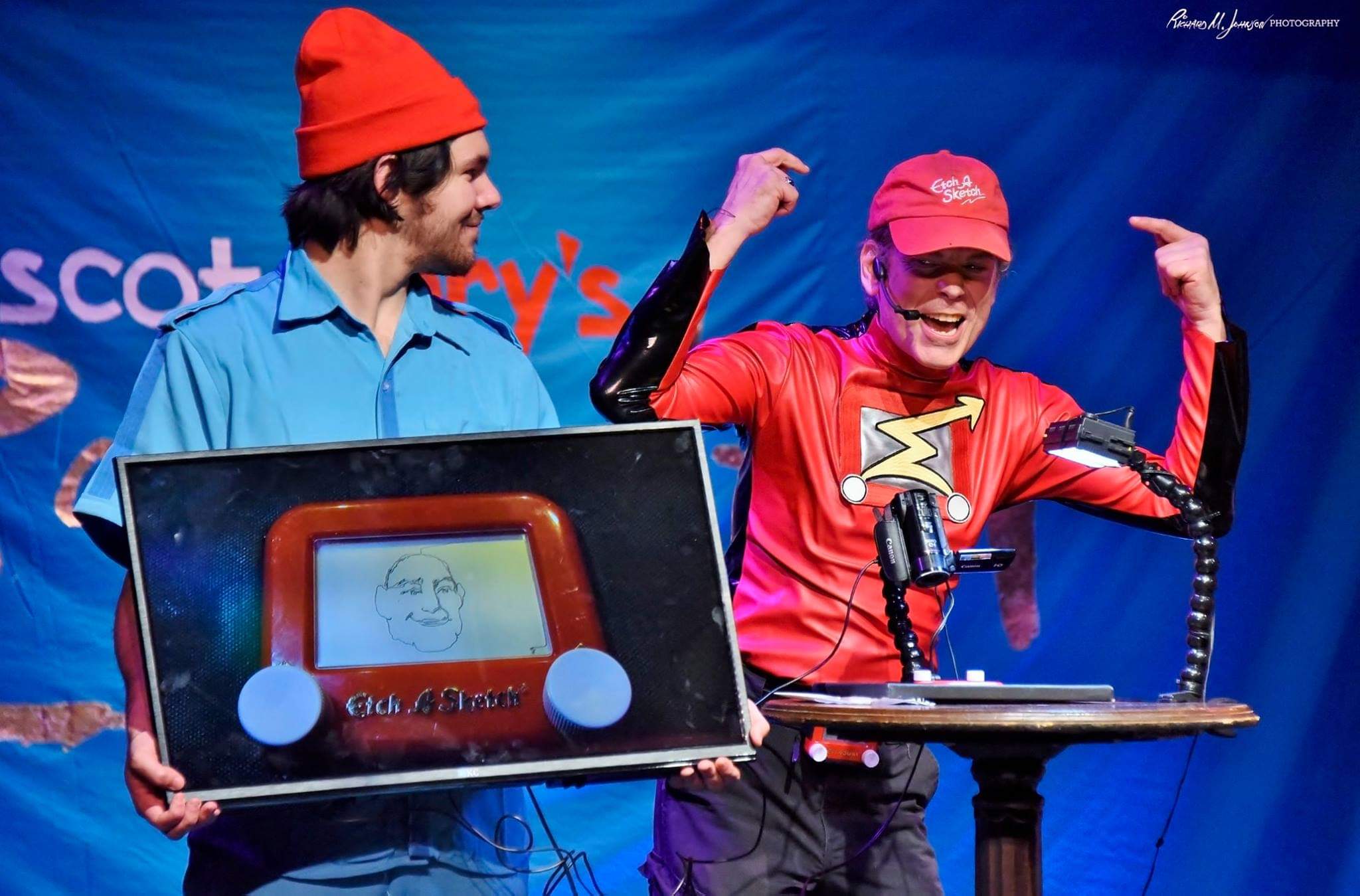 Two people on stage, one holding a large screen, the other wearing a red cap and shirt, smiling and flexing muscles.