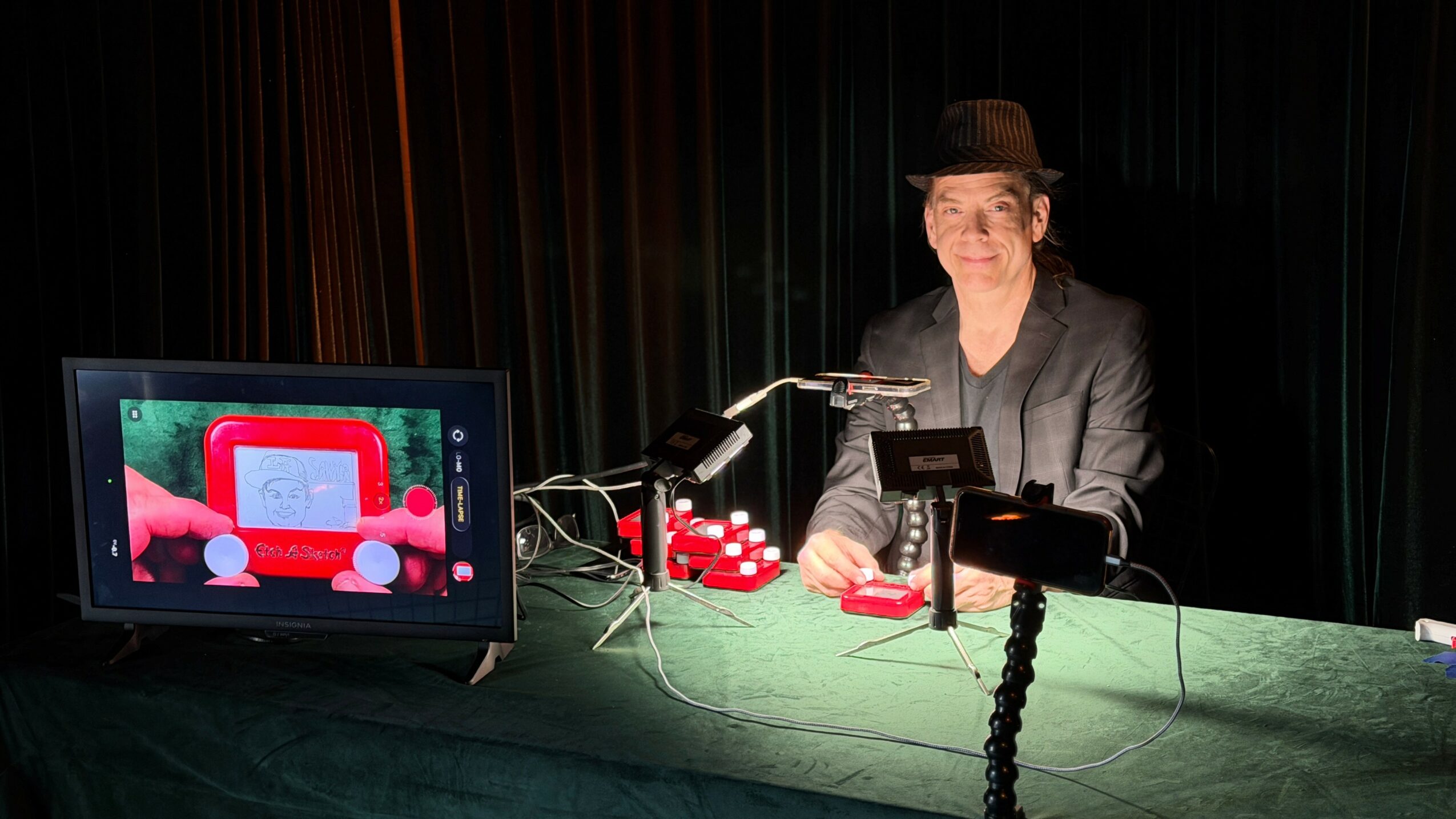 Person in a hat and glasses at a table with poker chips, a card shuffler, and a monitor, against a dark background.