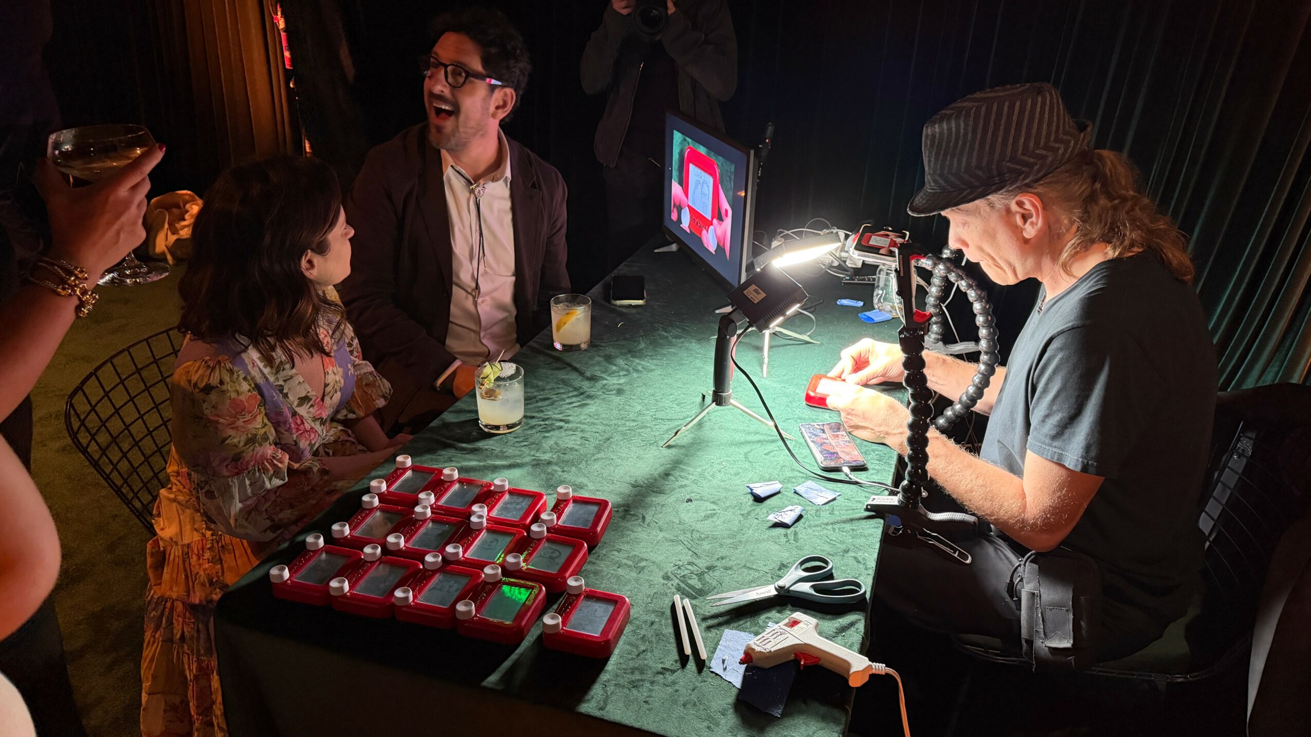 Person wearing a hat and glasses working with jewelry at a table with tools and jewelry displays, others watching nearby.