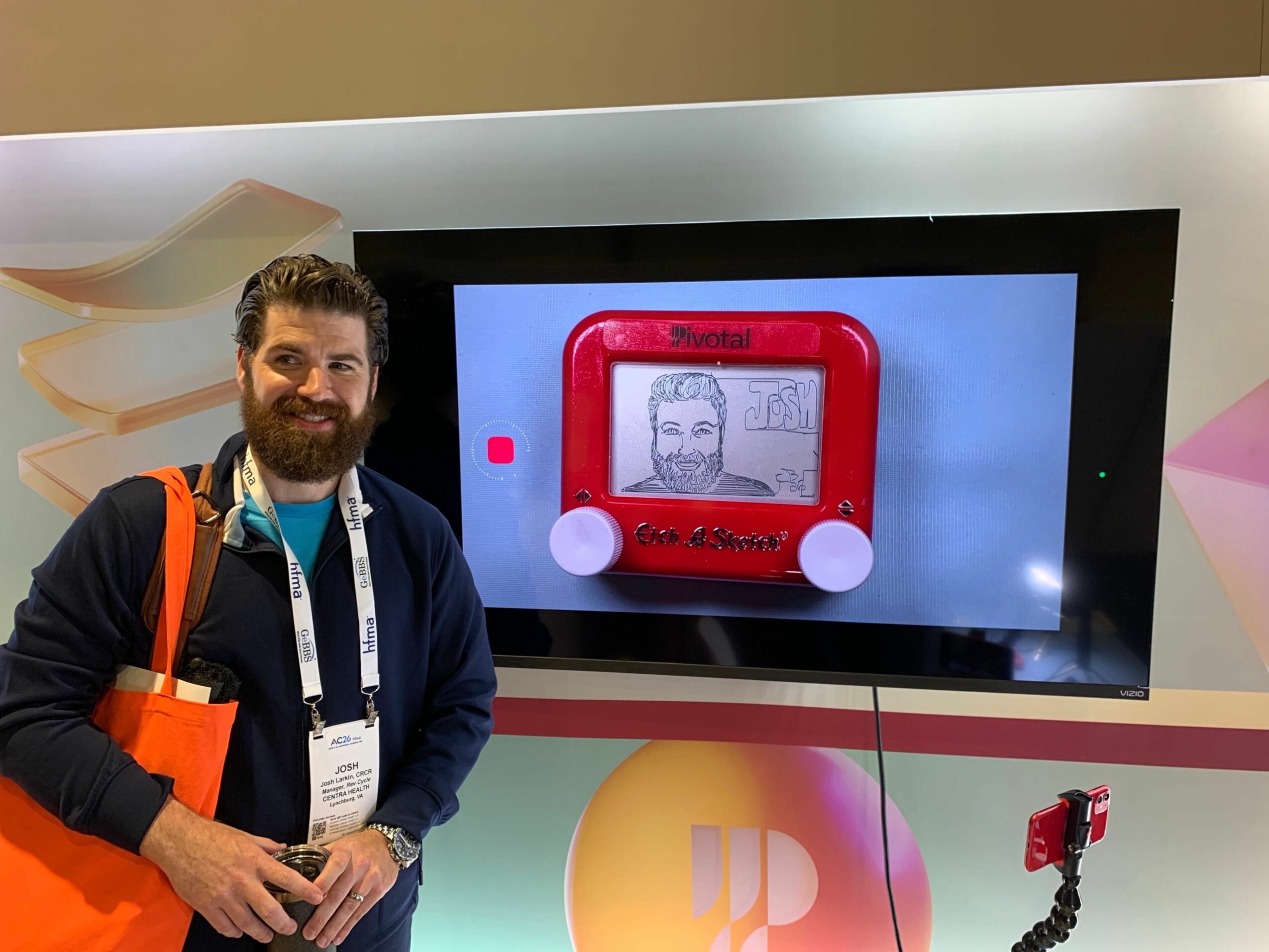 Person holding a device with a display showing a drawing, standing next to a large screen with a red frame, in an indoor setting.