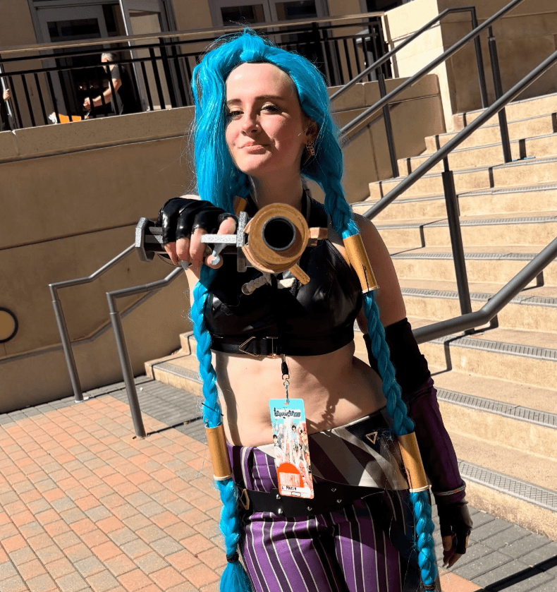 Person with bright blue hair taking a selfie outdoors on stairs, wearing a black top and purple striped pants.