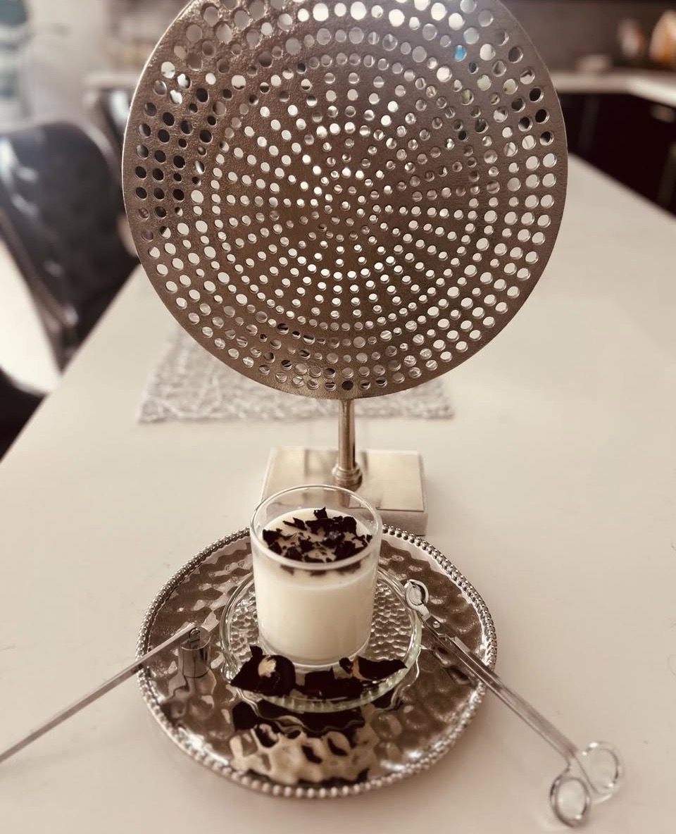 A metal strainer is positioned vertically behind a glass of layered dessert on a table, with a silver tray underneath.
