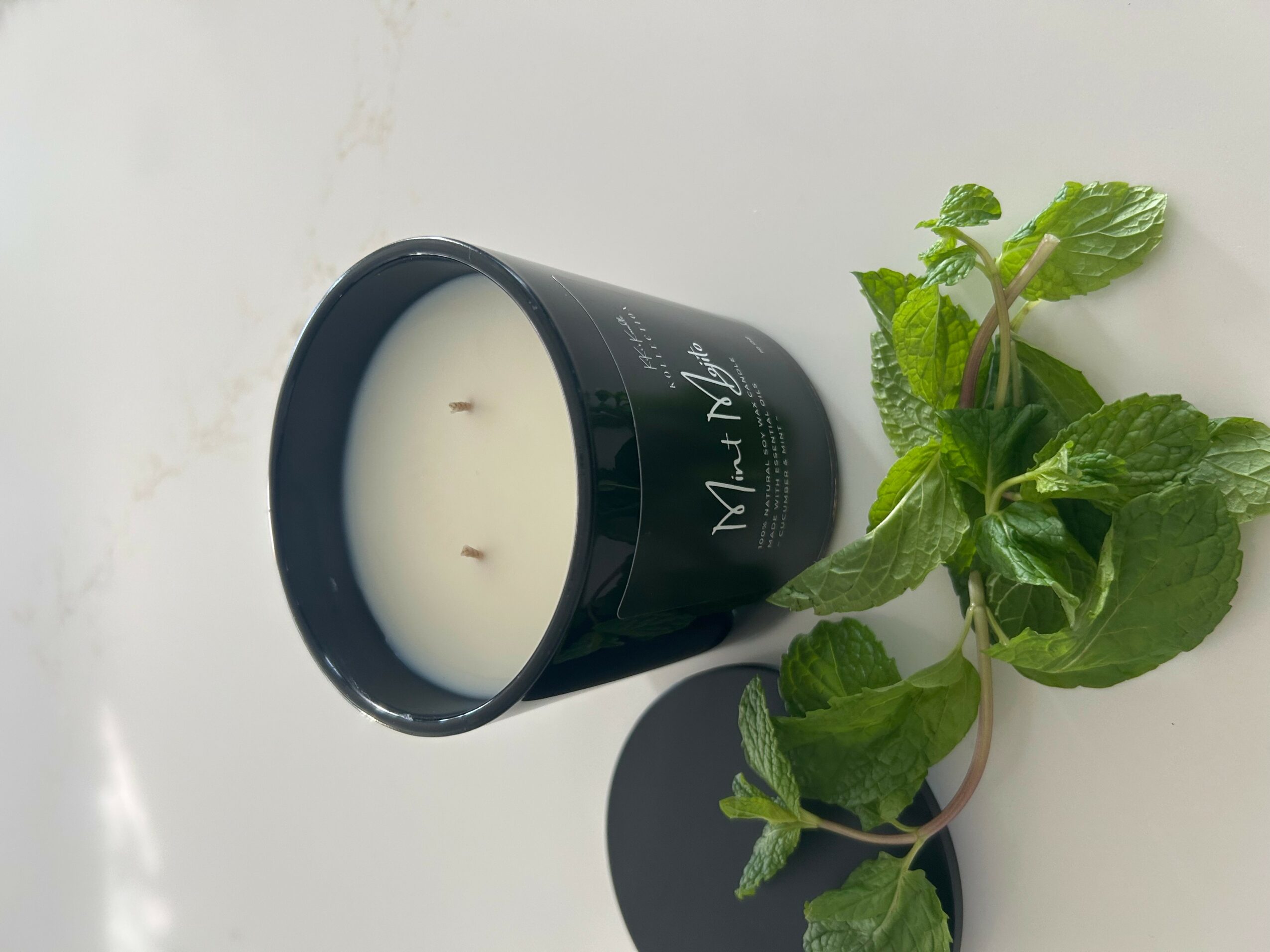 Black candle jar with white candle, mint leaves beside it, on a white surface.
