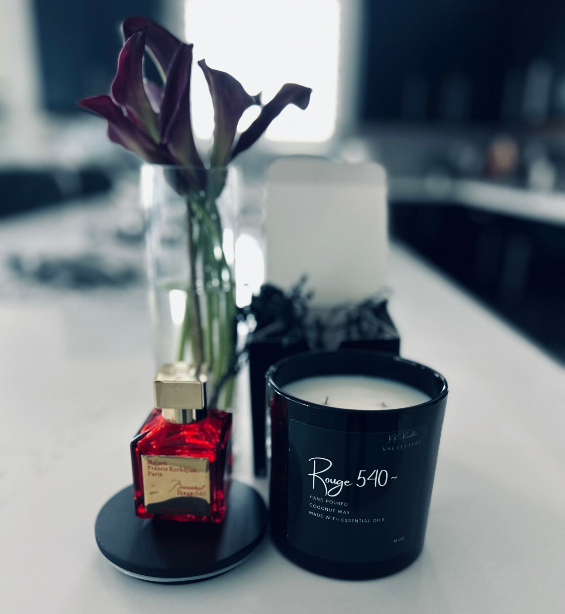 Vase with purple flowers, red perfume bottle, and black candle on a tray on a white surface.