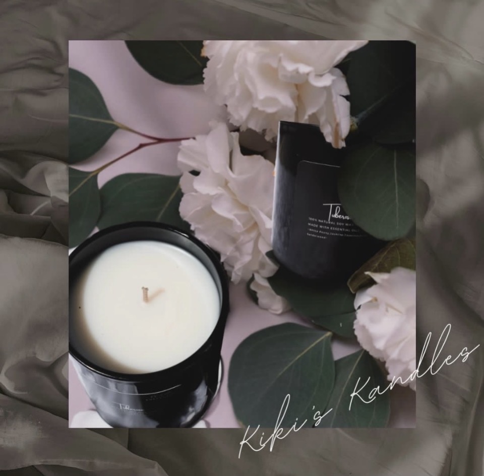 White candle in a black container surrounded by white flowers and green leaves, with a black box nearby, on a pink surface.