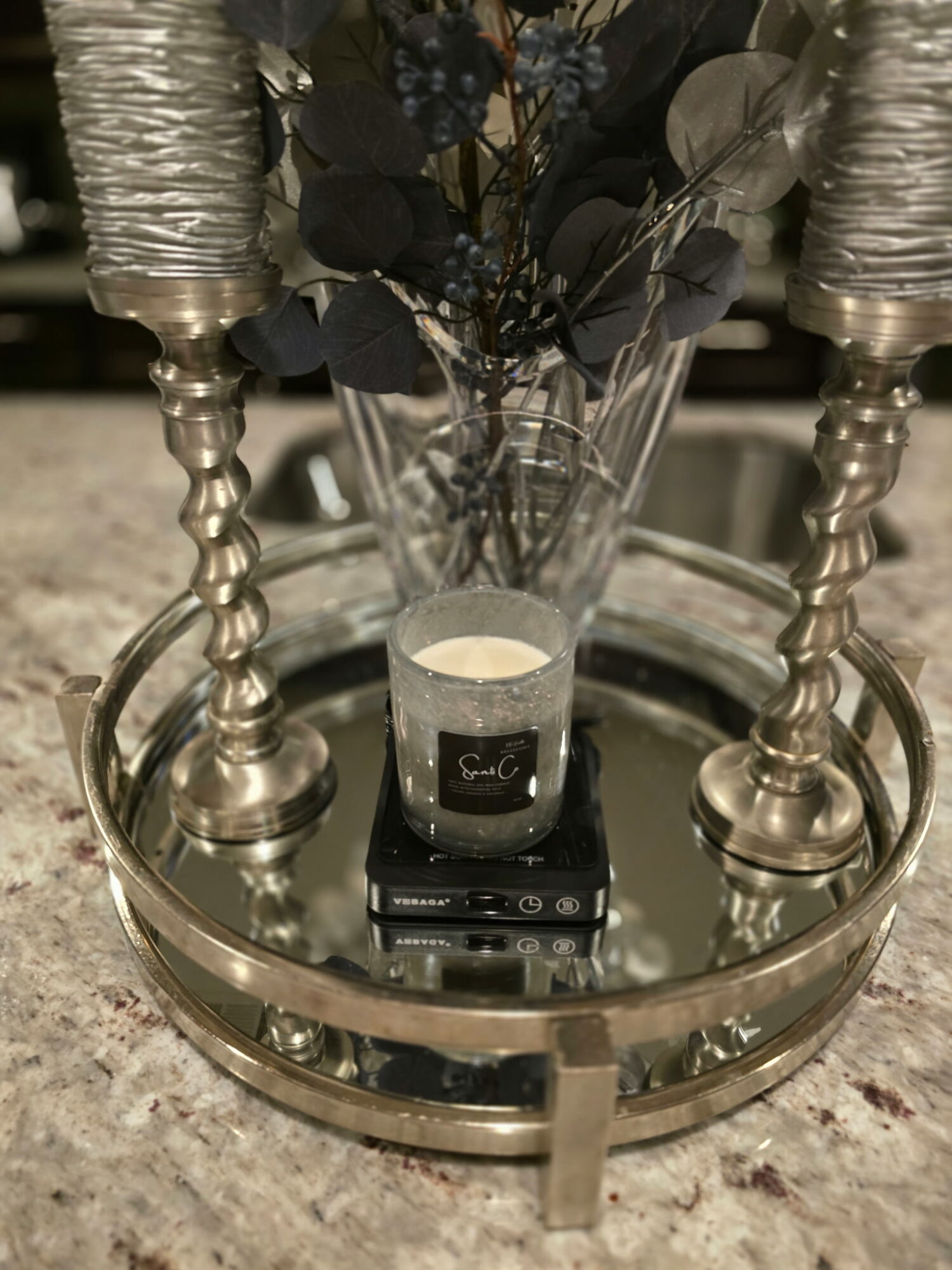 Decorative tray with candles, a glass vase with flowers, and a small black object on a countertop.
