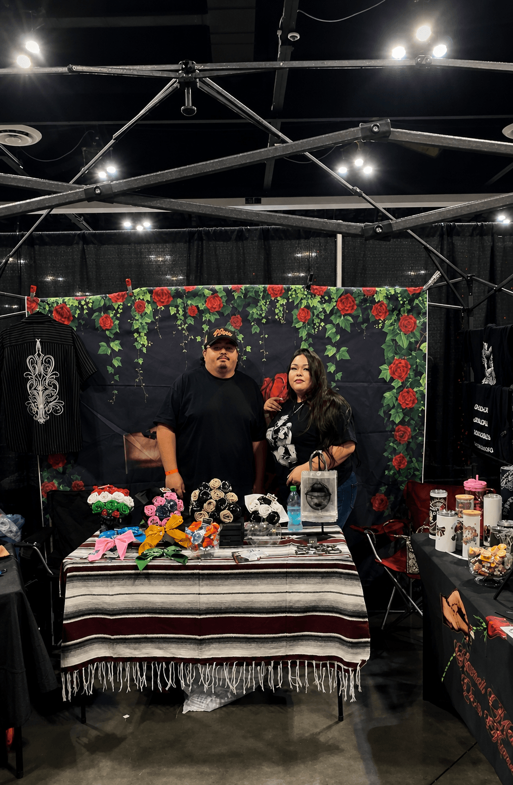 Two people stand behind a table with flowers and decorative items, in front of a black backdrop with red and green floral decorations.