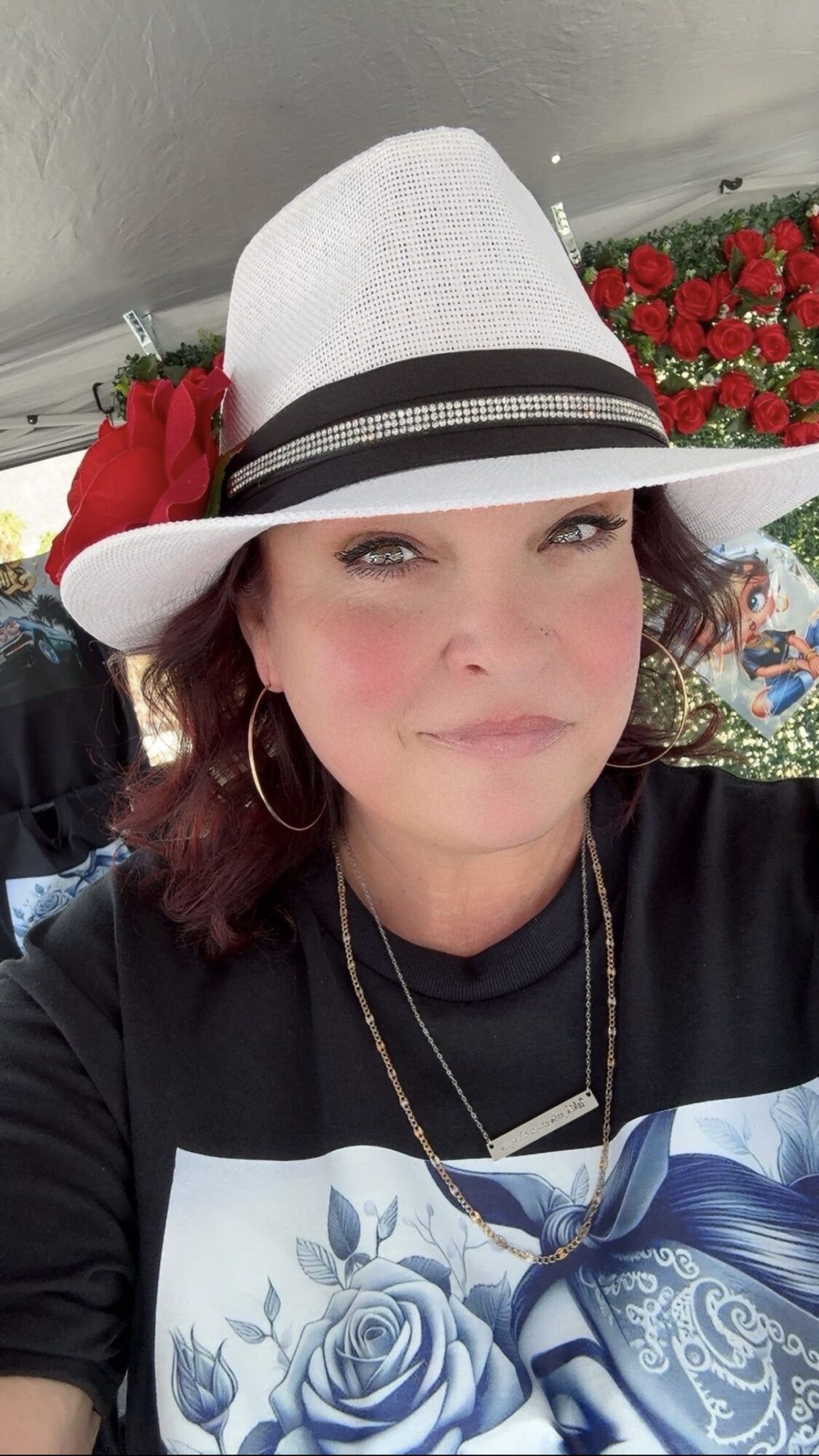 Woman wearing a white hat with black band, red flower, hoop earrings, and a black top with a graphic design, outdoors.