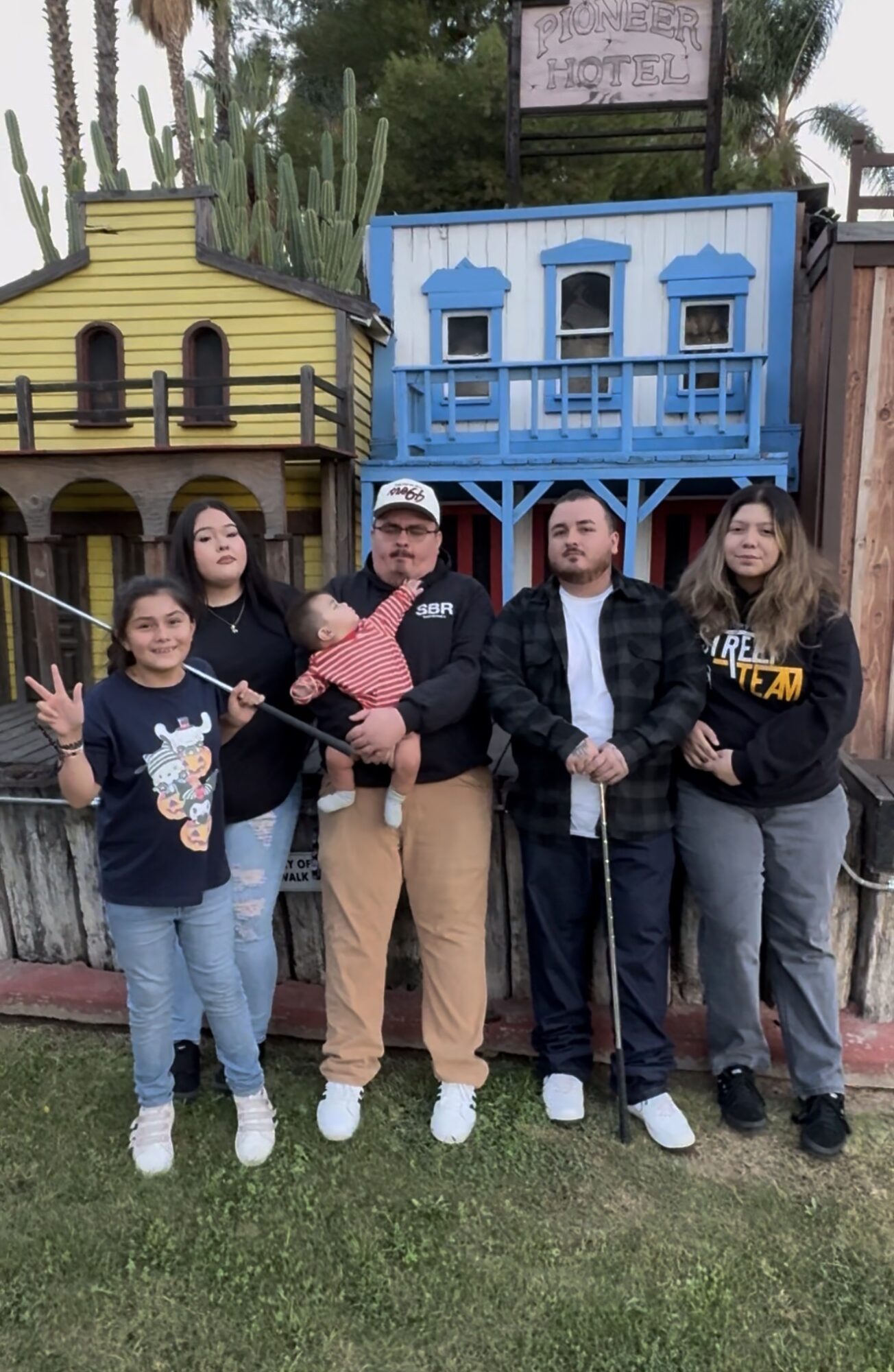Group of five people standing outdoors in front of colorful buildings, some holding objects, smiling, with grass and trees in background.