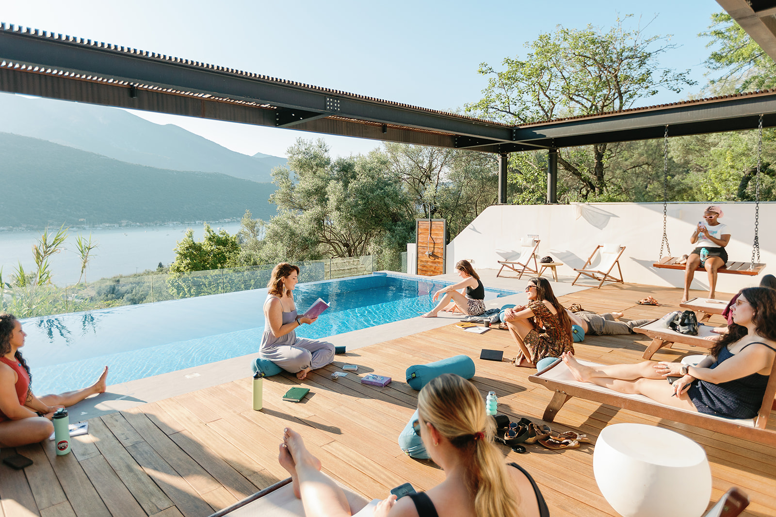 People relaxing by a poolside with lounge chairs, trees, and a lake in the background, under a clear sky.