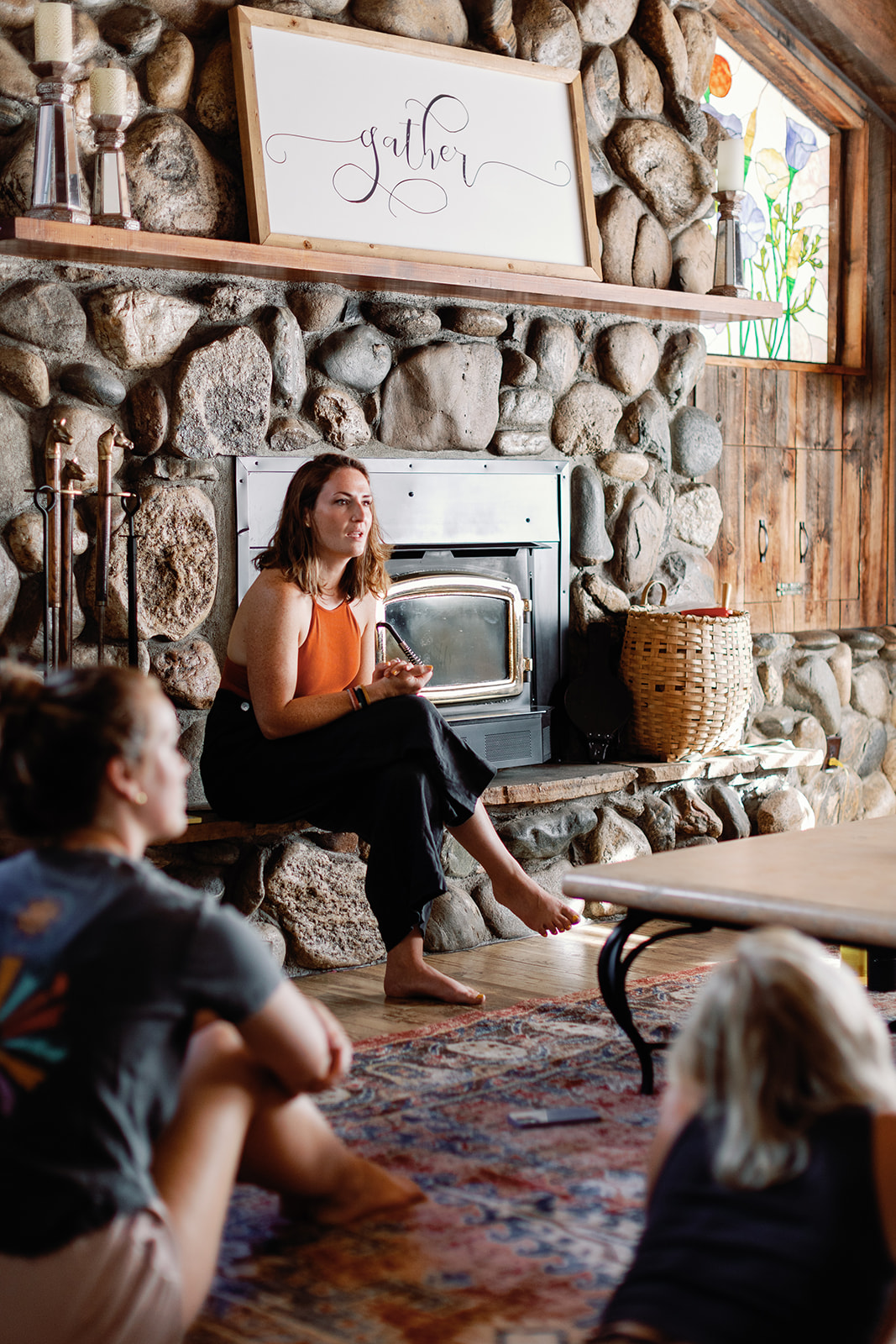 Woman sitting on a stone fireplace hearth, holding a mug, with children sitting on a rug in a cozy room.