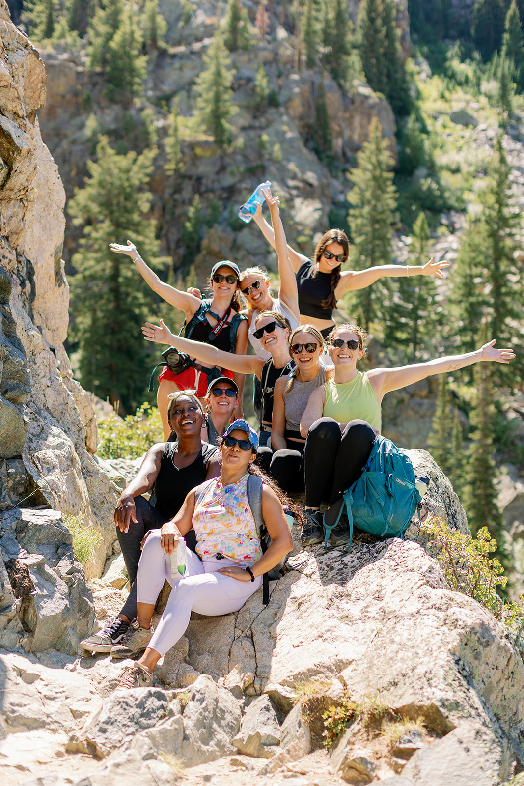 Group of seven people hiking on rocky terrain in a forested mountain area, some with arms outstretched, wearing outdoor clothing and sunglasses.