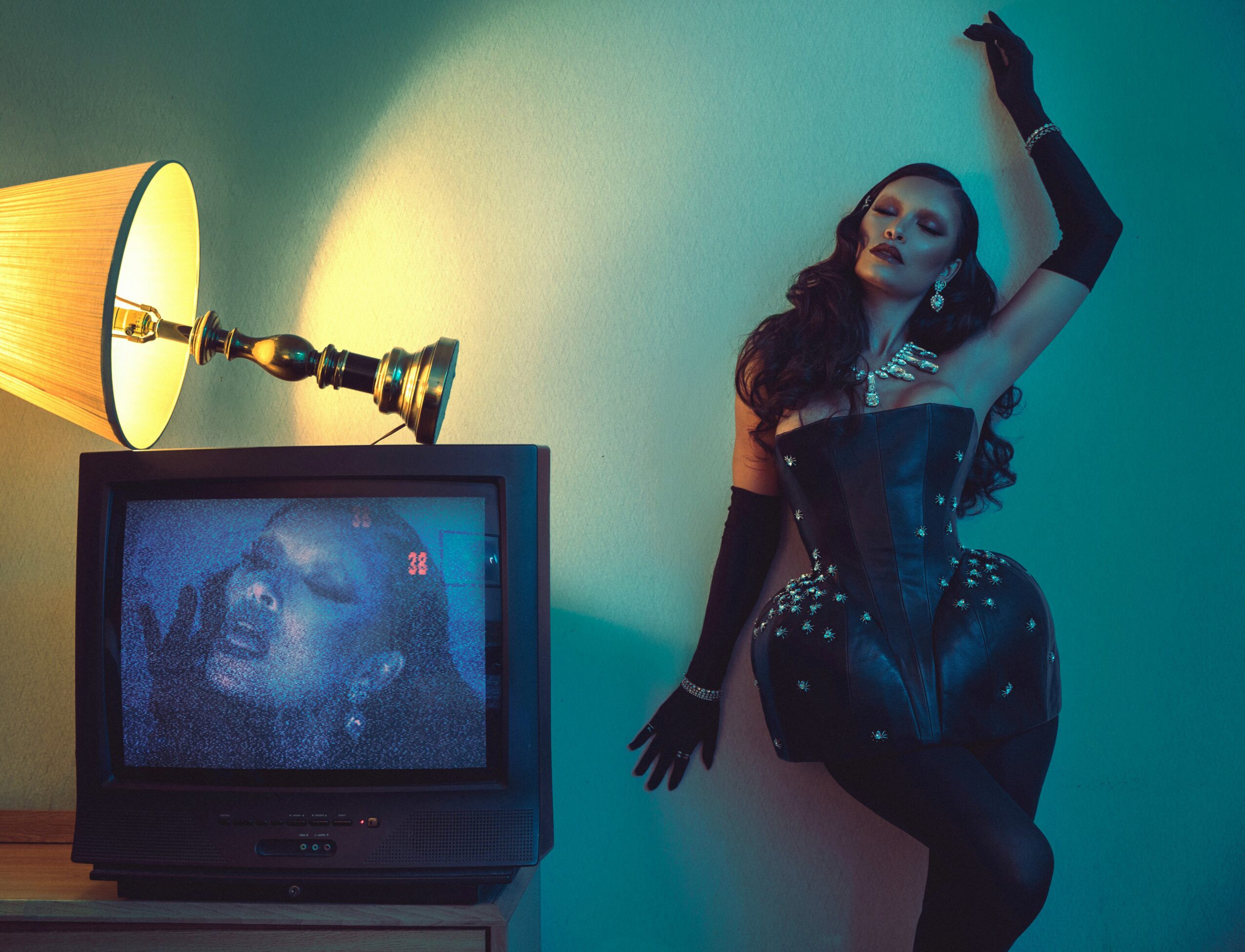 Woman in black outfit and gloves poses next to a TV showing a close-up of her face, with a lamp nearby.