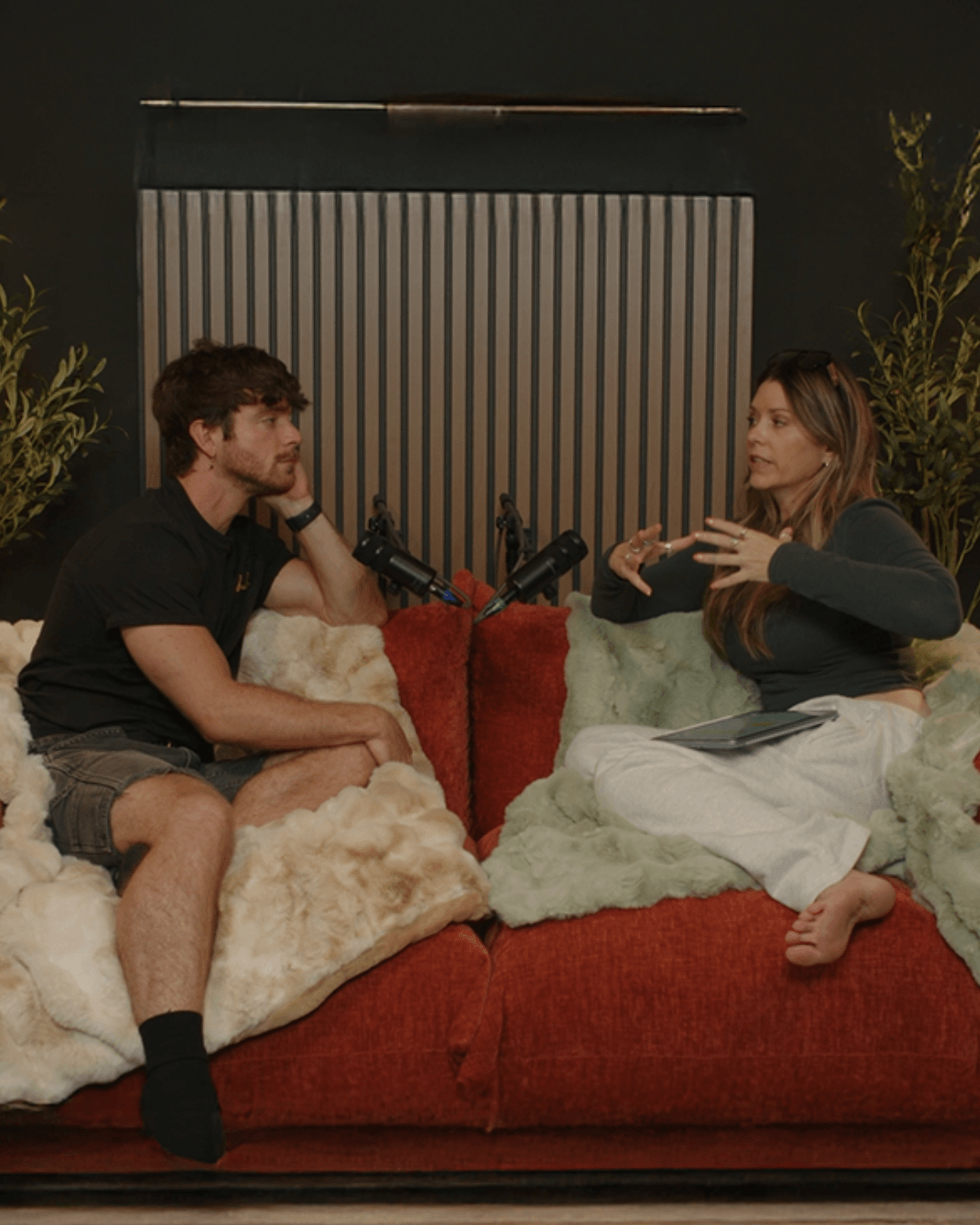 Two people sitting on a couch, engaged in conversation, with a microphone between them, in a dimly lit room.