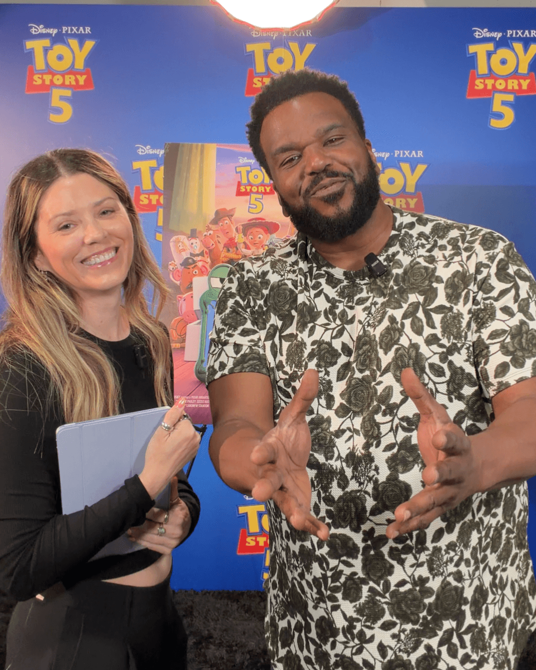 Two people smiling in front of a Toy Story 5 poster, one holding a notebook, the other making a gesture with hands.
