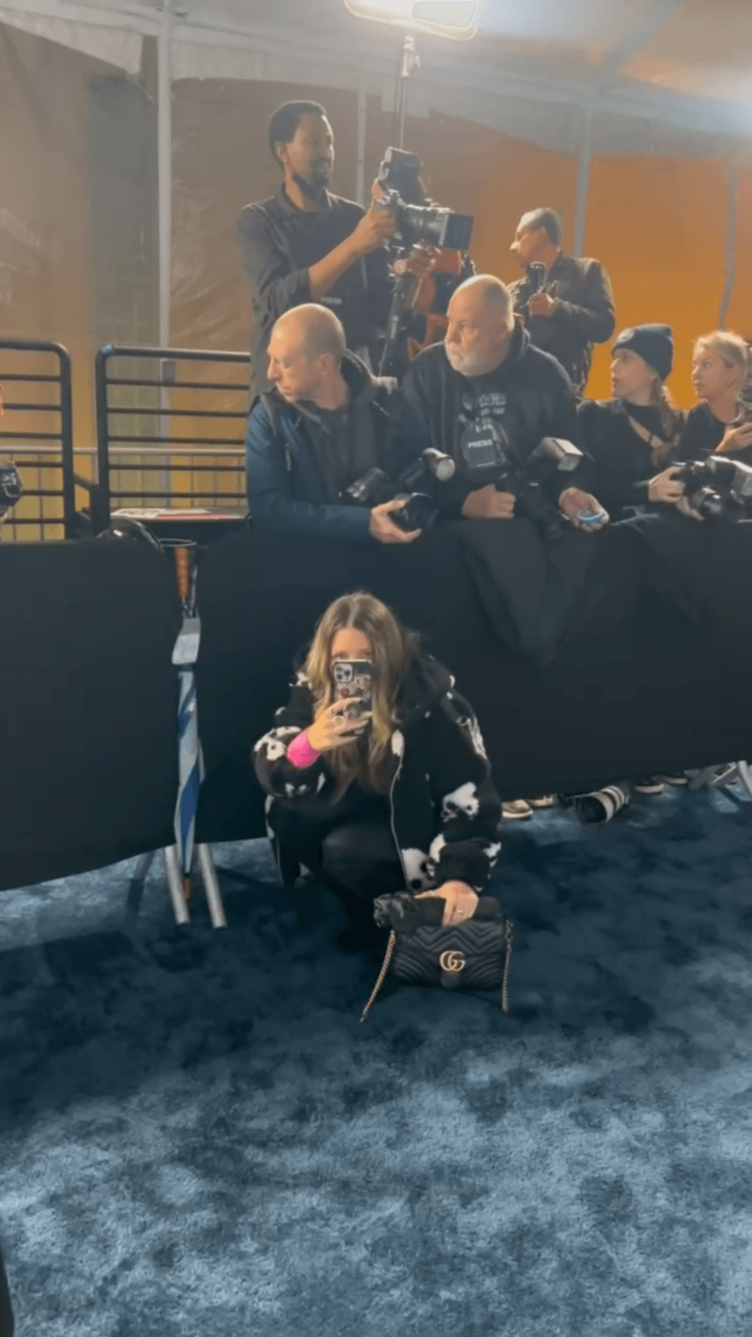 A woman with long hair and a face mask kneels on a blue carpet, holding a camera, with a group of people behind her.