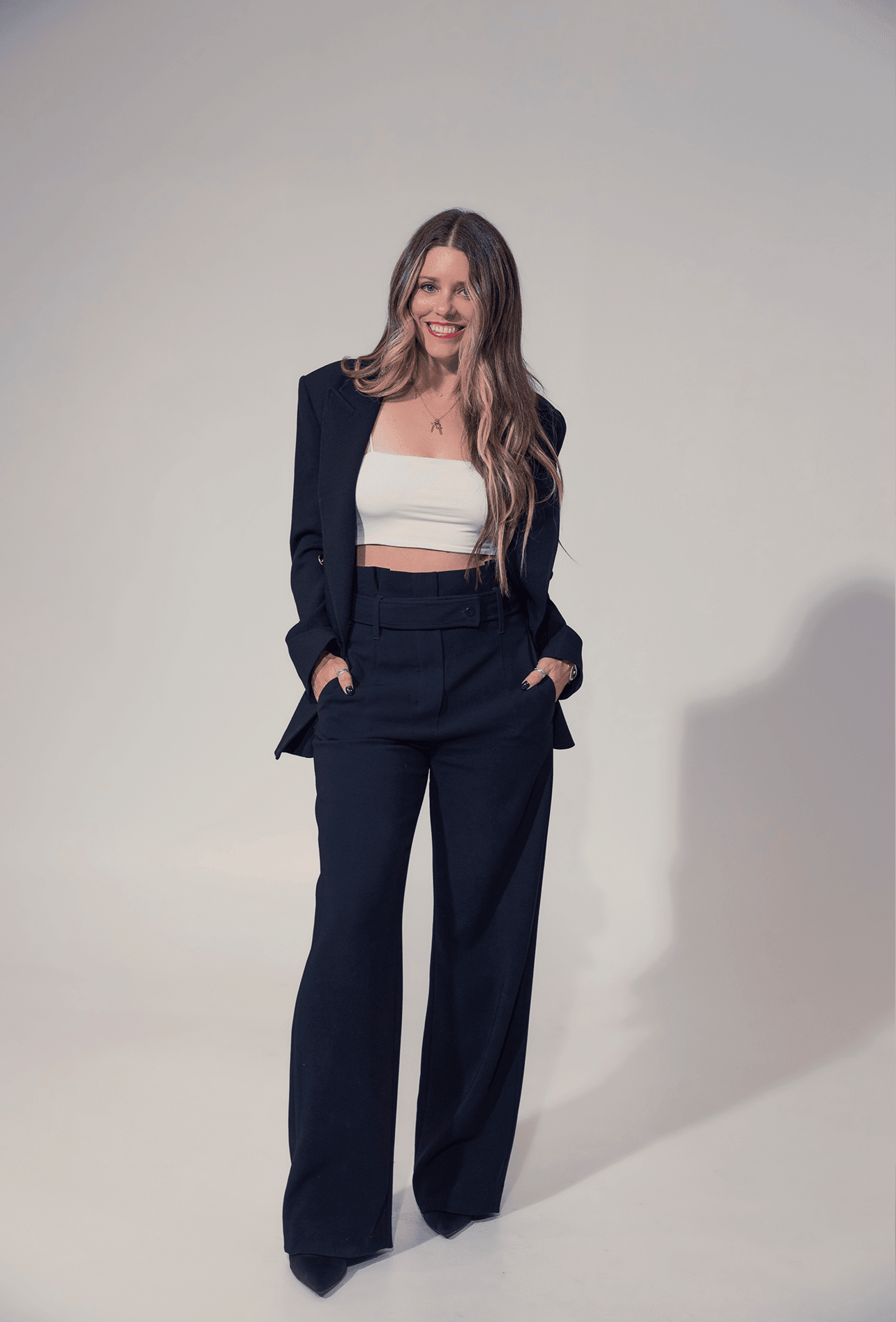 Woman with long hair in a black suit and white top, standing with hands in pockets, smiling, against a plain background.